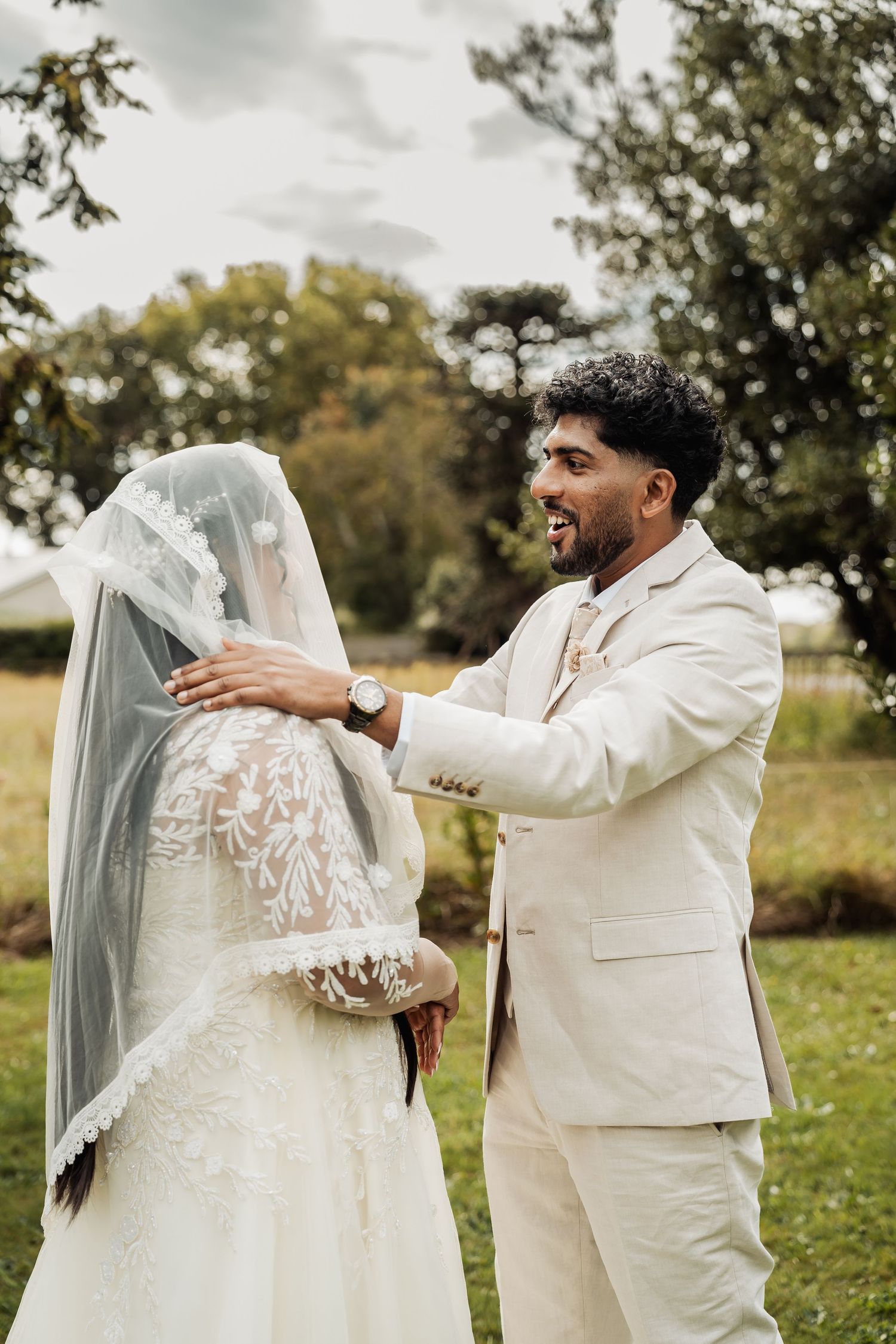 Groom in white suit smiling at bride in lace wedding dress and veil during outdoor first look ceremony.