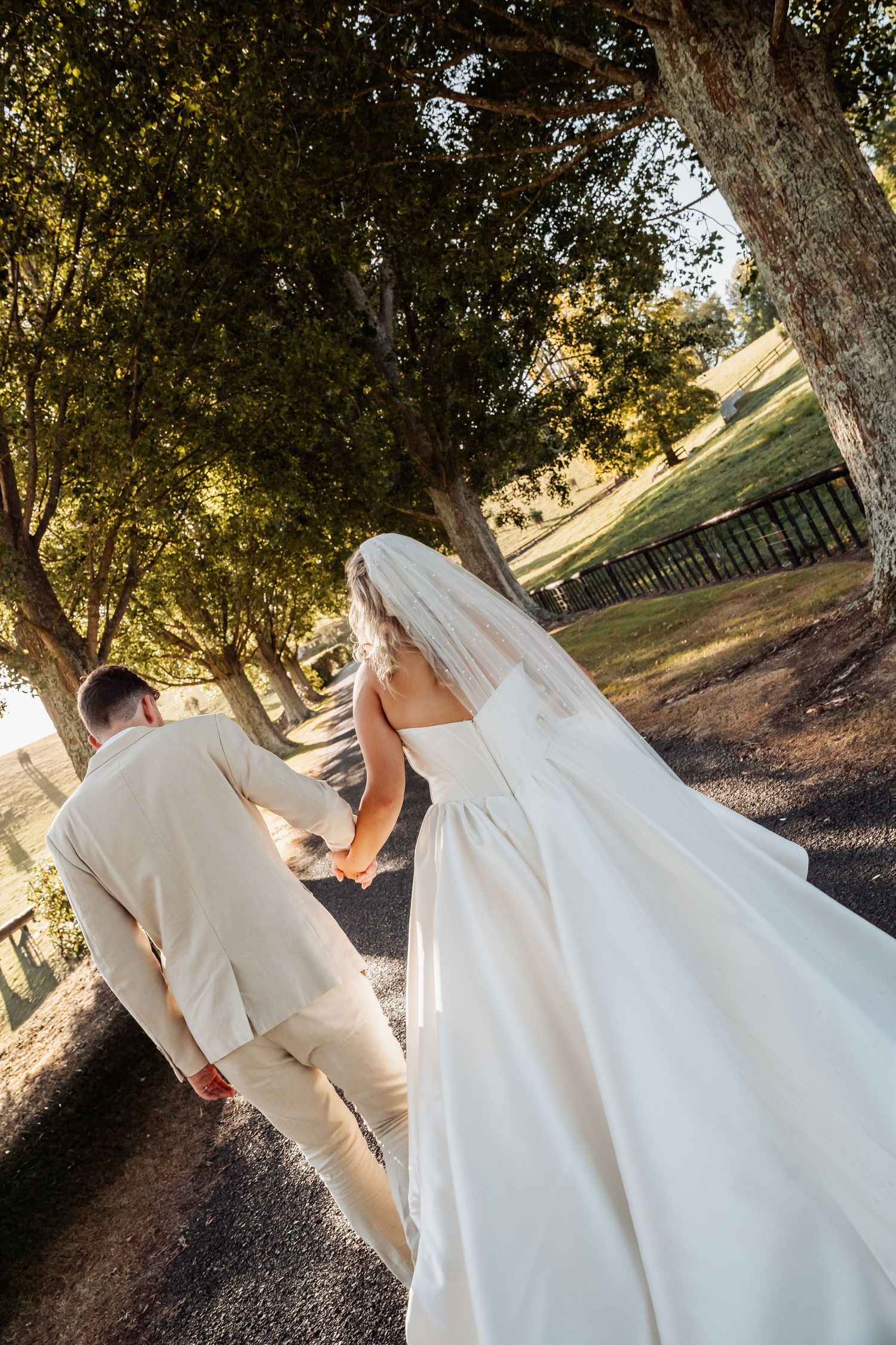 Bride and groom walk hand-in-hand down a tree-lined path on their wedding day.