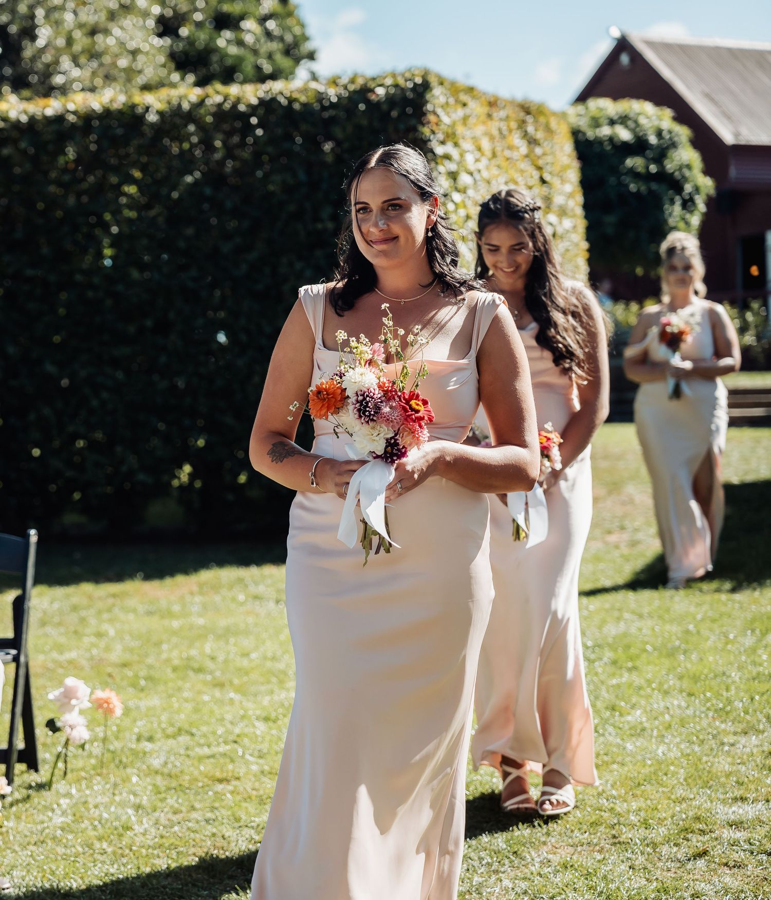 Bridesmaids in champagne gowns walking down outdoor aisle carrying colorful bouquets.