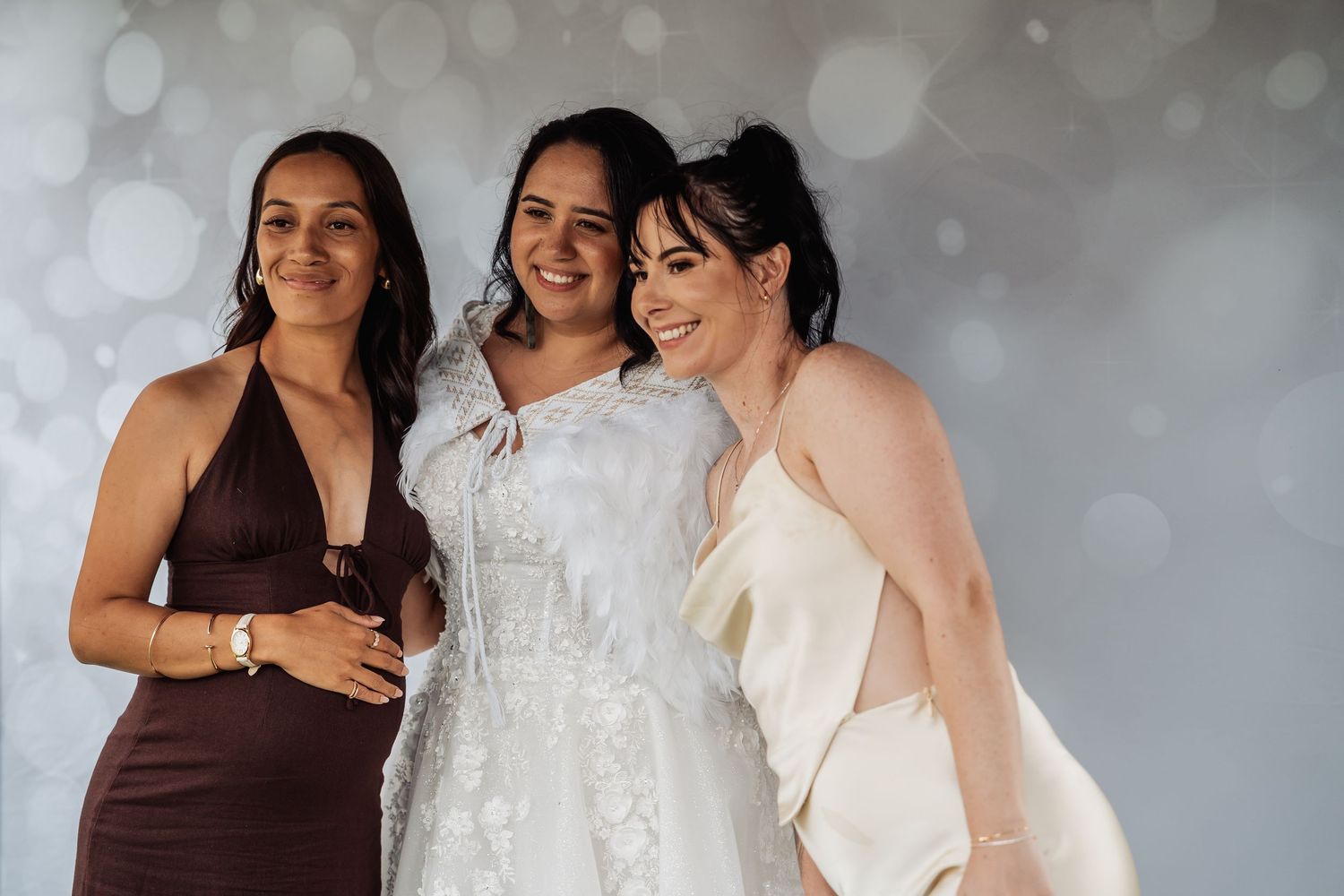 Bride poses with two friends in front of a bokeh backdrop at wedding celebration.