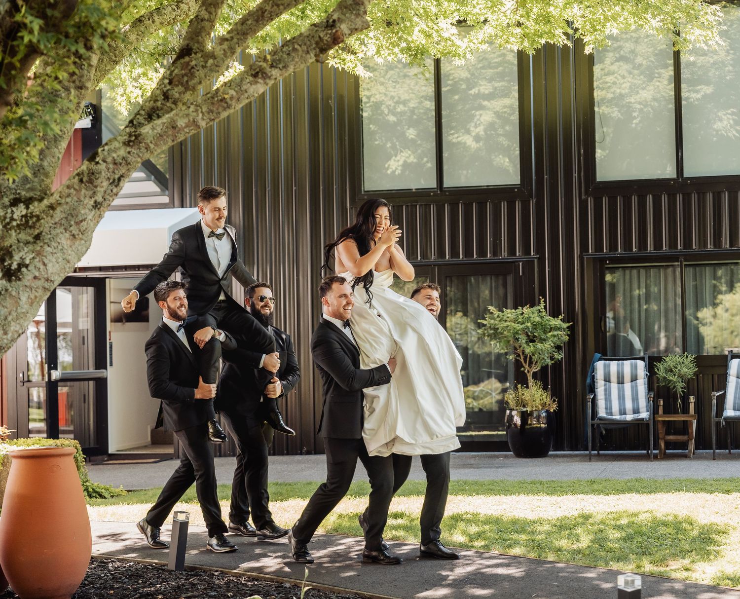 Groomsmen playfully lift the bride outdoors near a modern building on the wedding day.