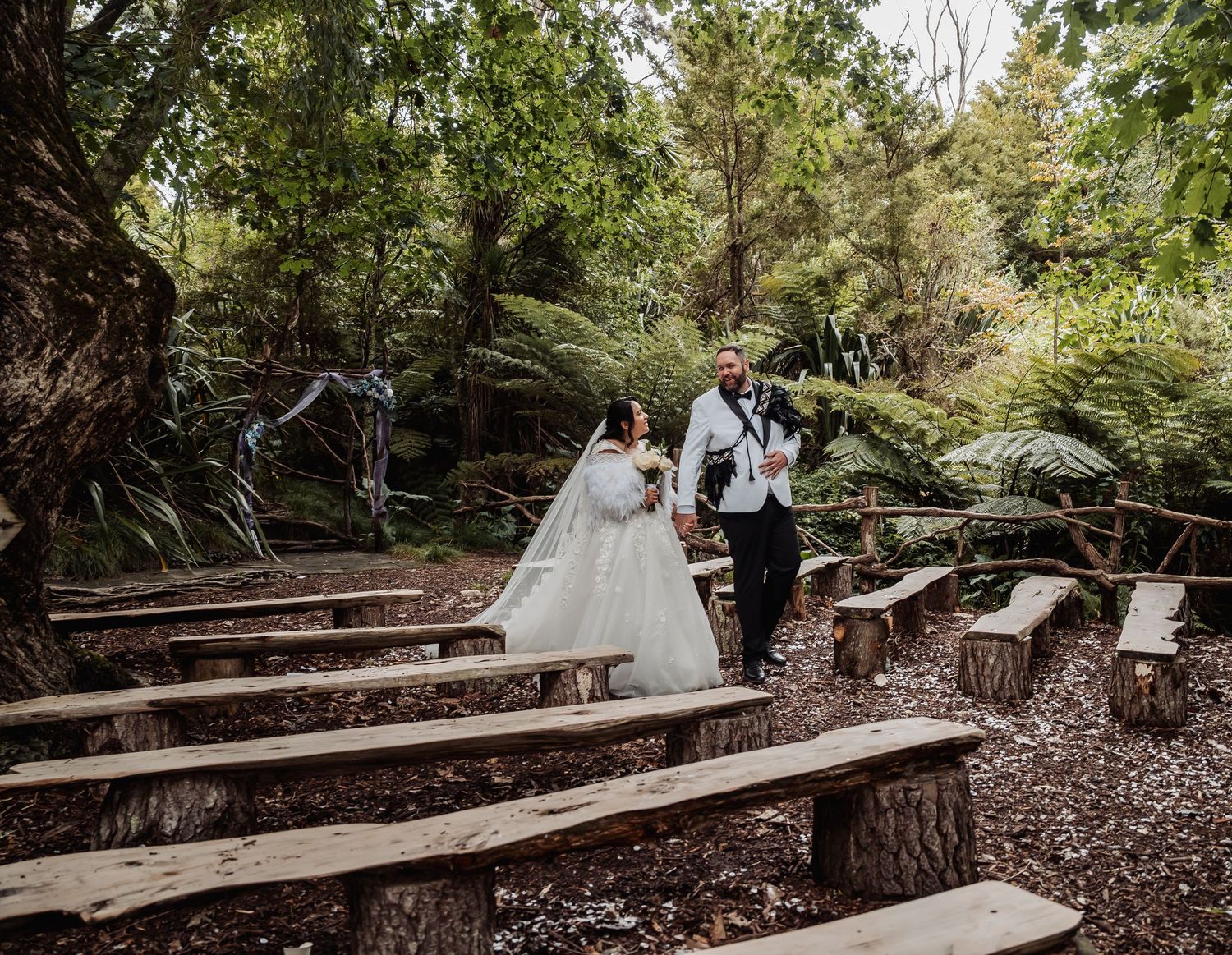 Newlywed couple walks through rustic woodland ceremony space with log bench seating.