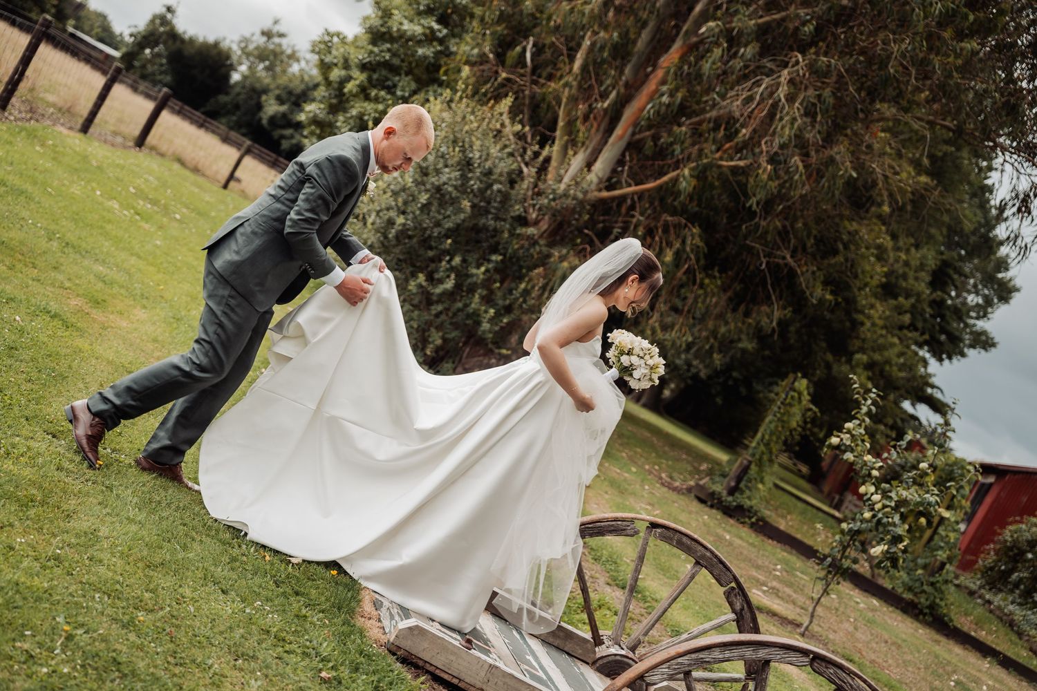 Groom helps lift bride's white wedding gown near a rustic wooden wagon in garden.