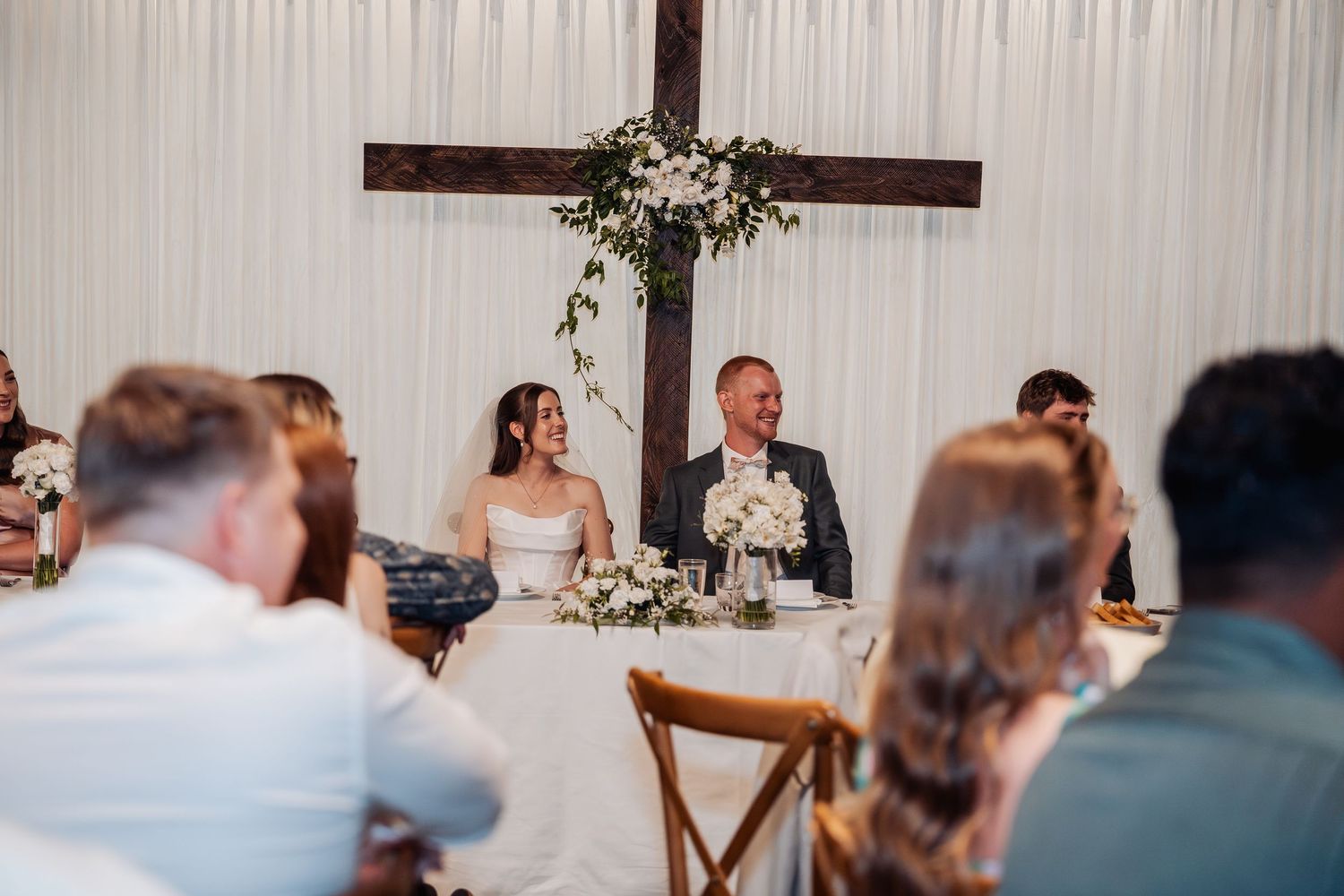 Bride and groom smile at sweetheart table with wooden cross backdrop during elegant wedding reception.