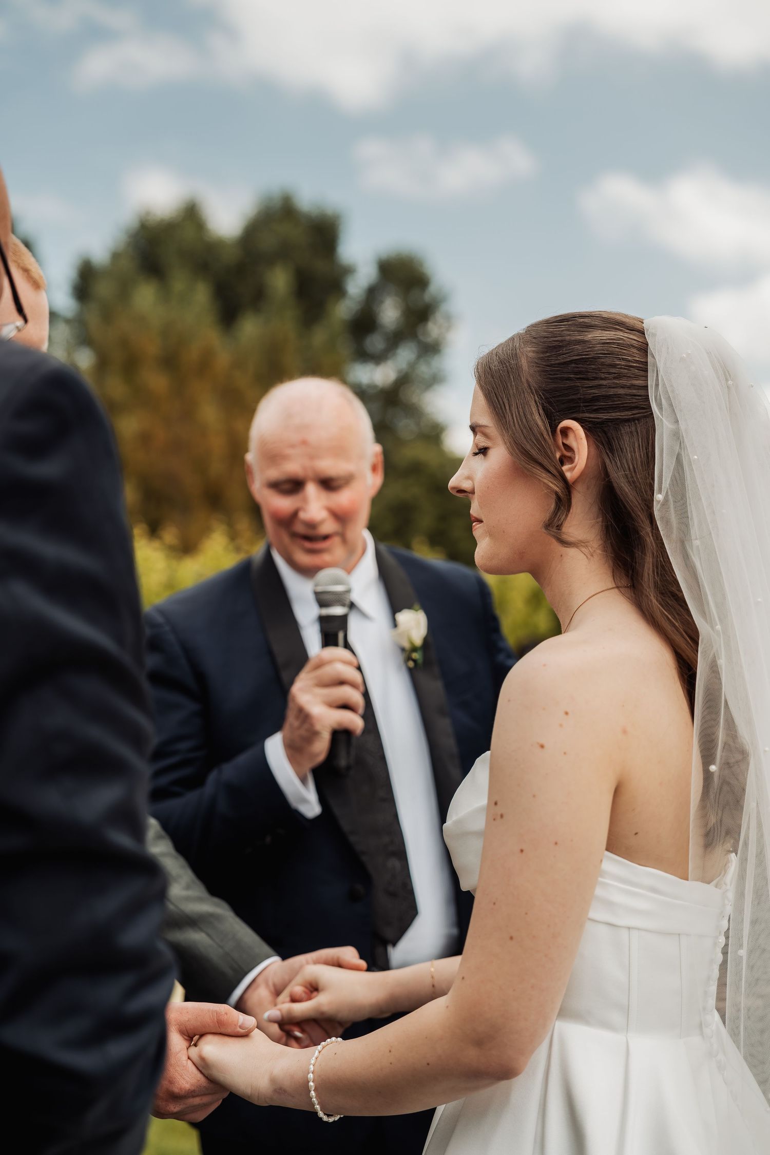 Bride in white strapless gown holds grooms hands during outdoor wedding ceremony officiated by bald officiant.