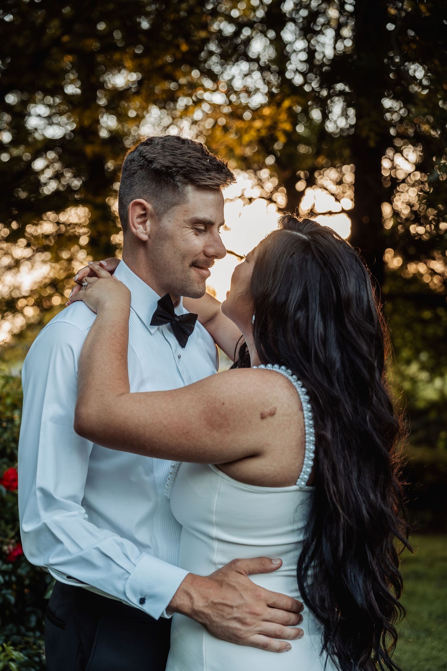 Bride and groom embracing and smiling at each other in golden sunset light among trees.