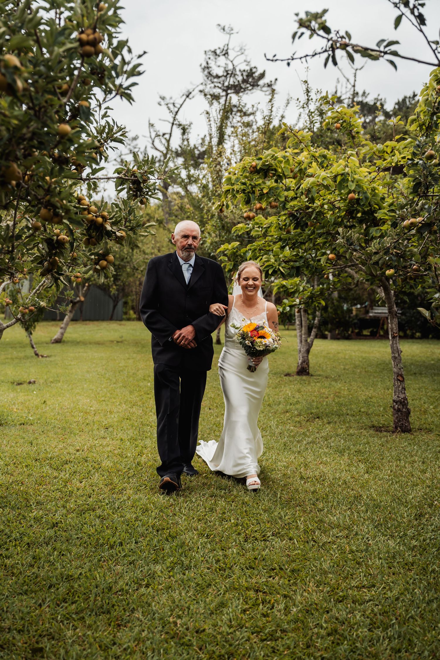 Father escorts bride in white gown through fruit tree orchard on wedding day.