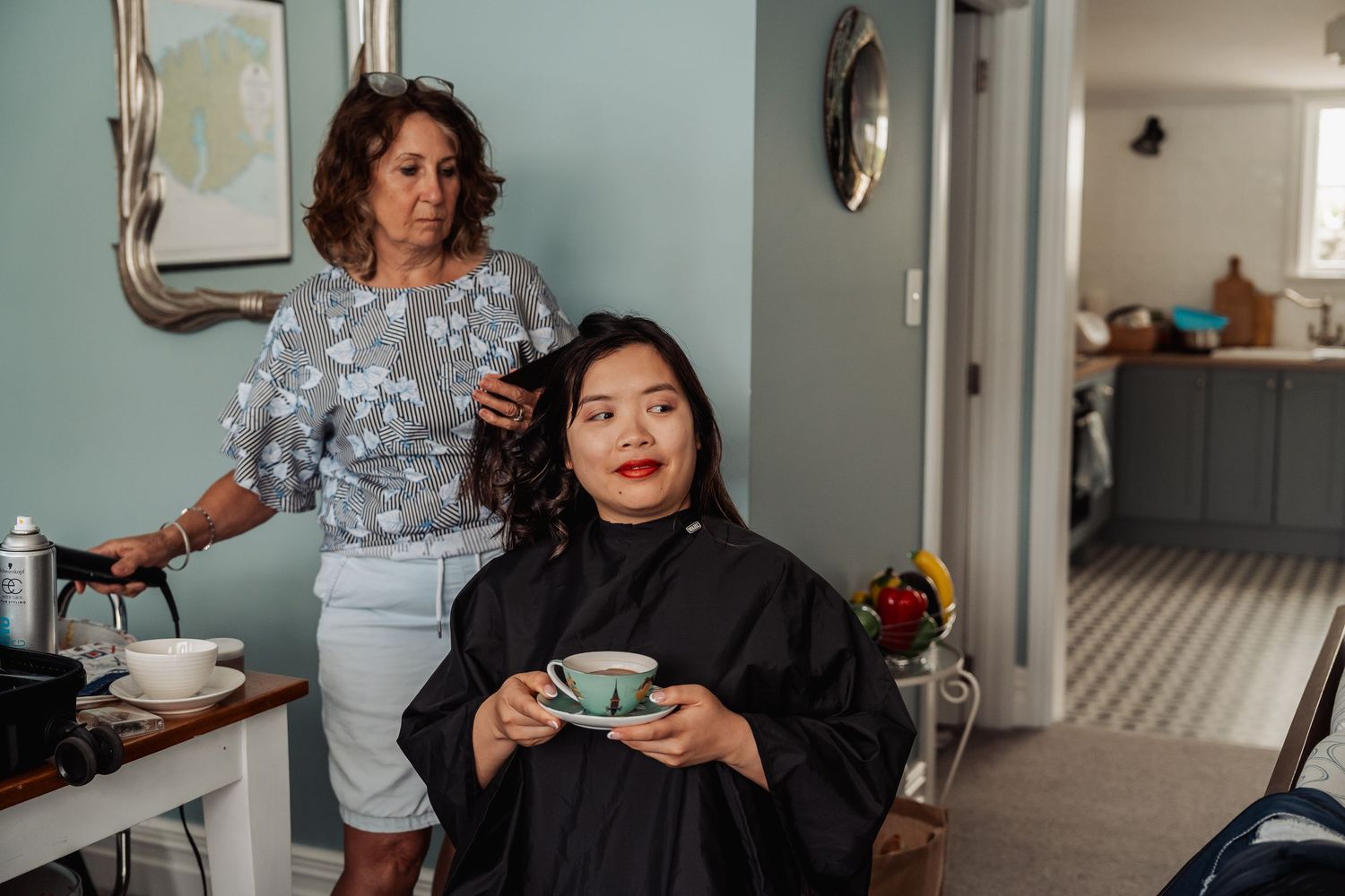 A hairstylist curls an Asian bride's hair while she relaxes holding a teal teacup during wedding preparations.