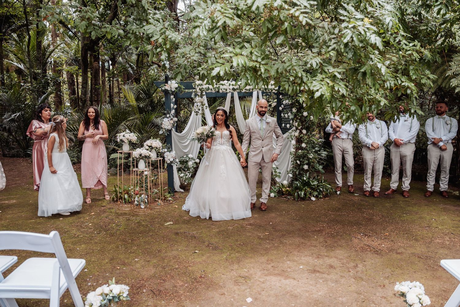 Bride and groom celebrate under floral arch with wedding party in lush garden ceremony setting.