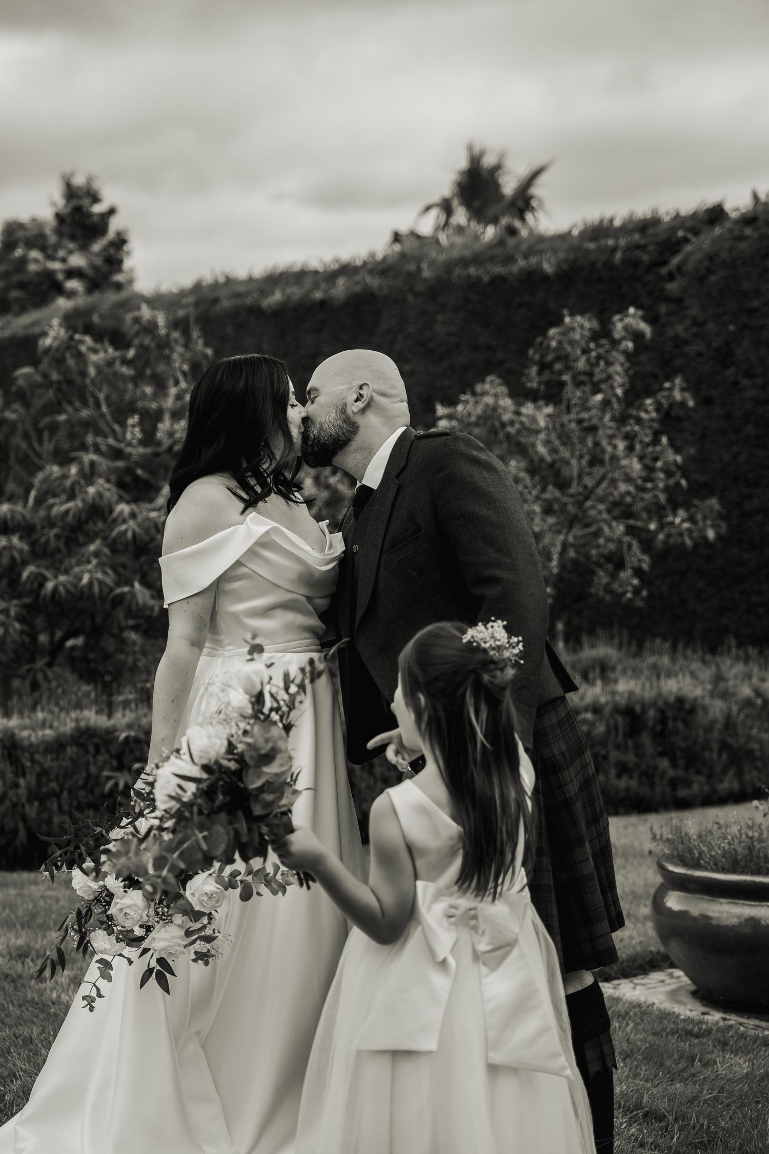 Black and white photo of bride and groom kissing with flower girl beside them outdoors.