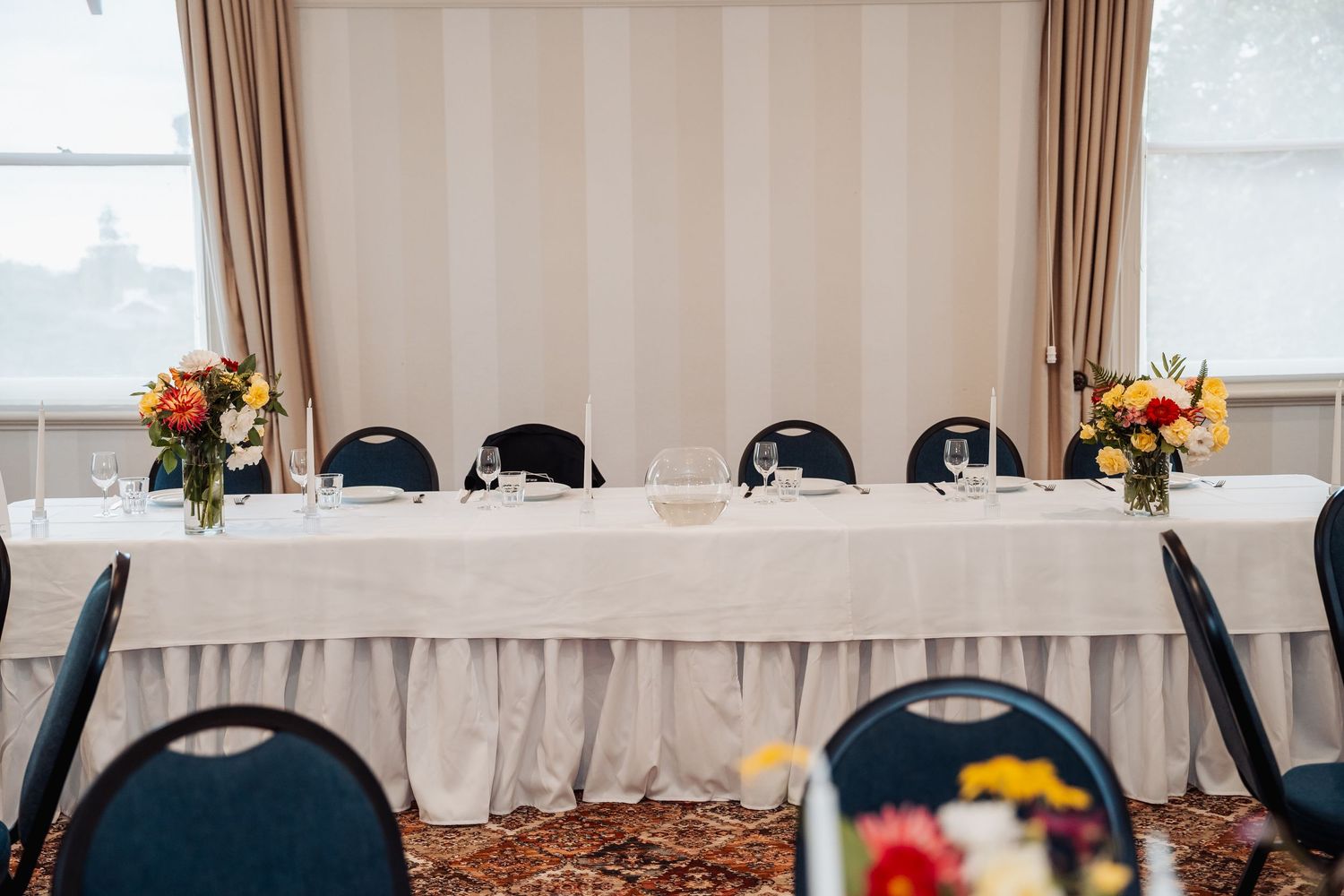 Elegant wedding head table with white linen, floral centerpieces of red and yellow flowers, and black chairs in a banquet hall.