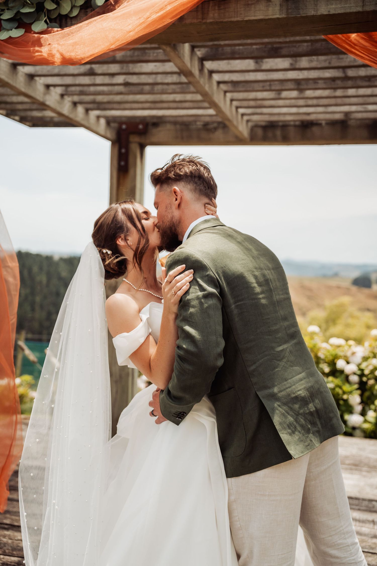 Bride and groom share first kiss at outdoor wedding ceremony with scenic hillside backdrop.