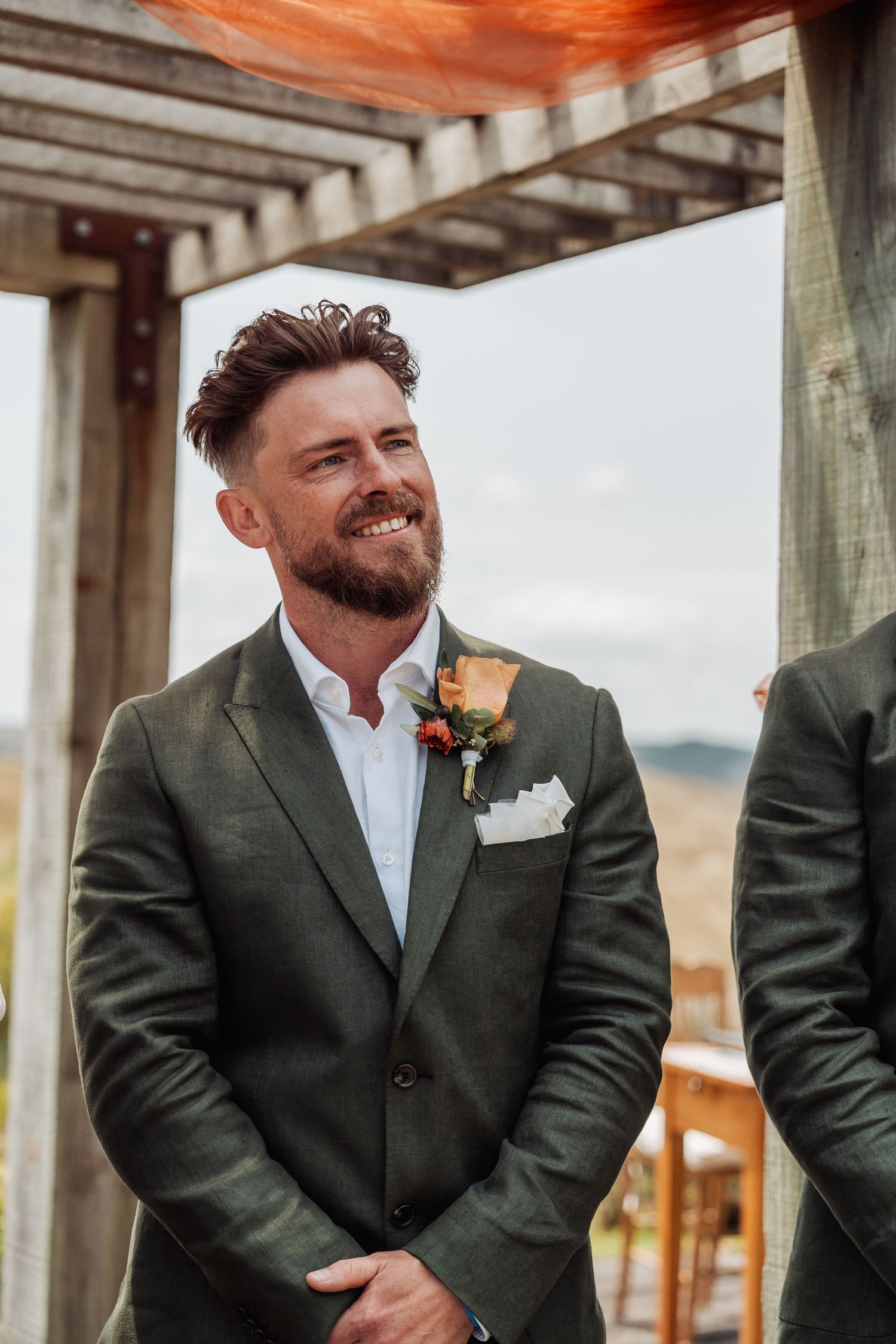 Smiling groom in dark green suit with floral boutonniere stands under wooden pergola at wedding ceremony.