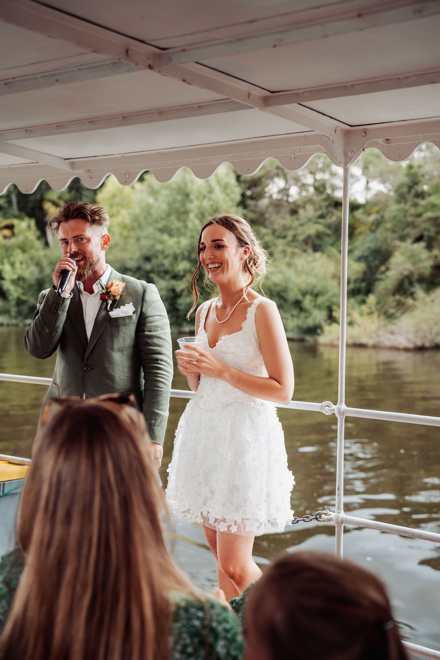 Bride in short white dress and groom in green suit laughing on a boat with guests during reception.