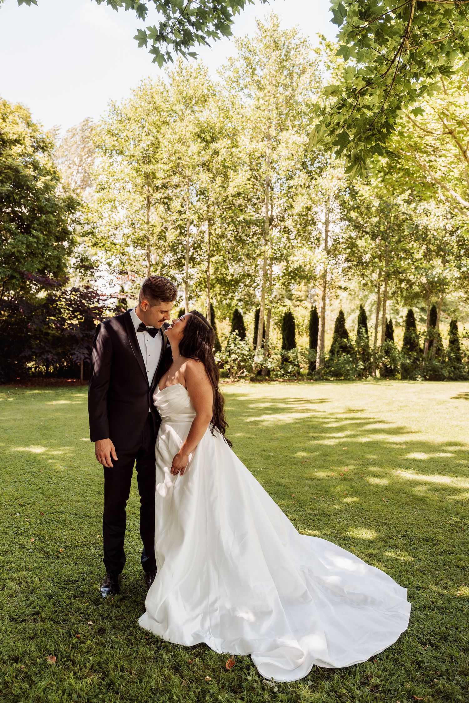 Bride in white gown and groom in black tuxedo share a romantic moment on lush green lawn surrounded by tall trees.
