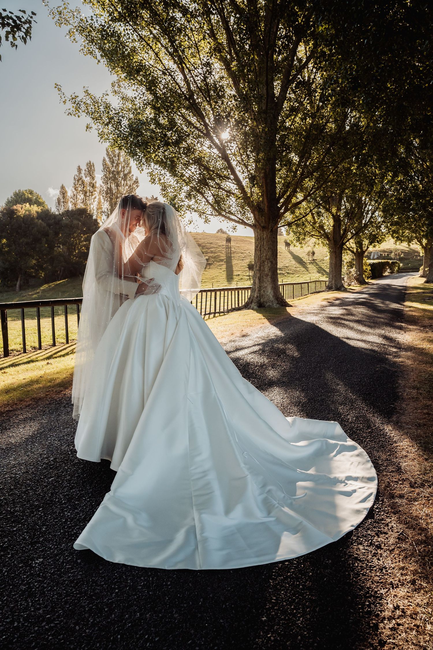 Bride and groom kissing under veil on tree-lined path, bride in elegant white gown with long train, golden sunlight.