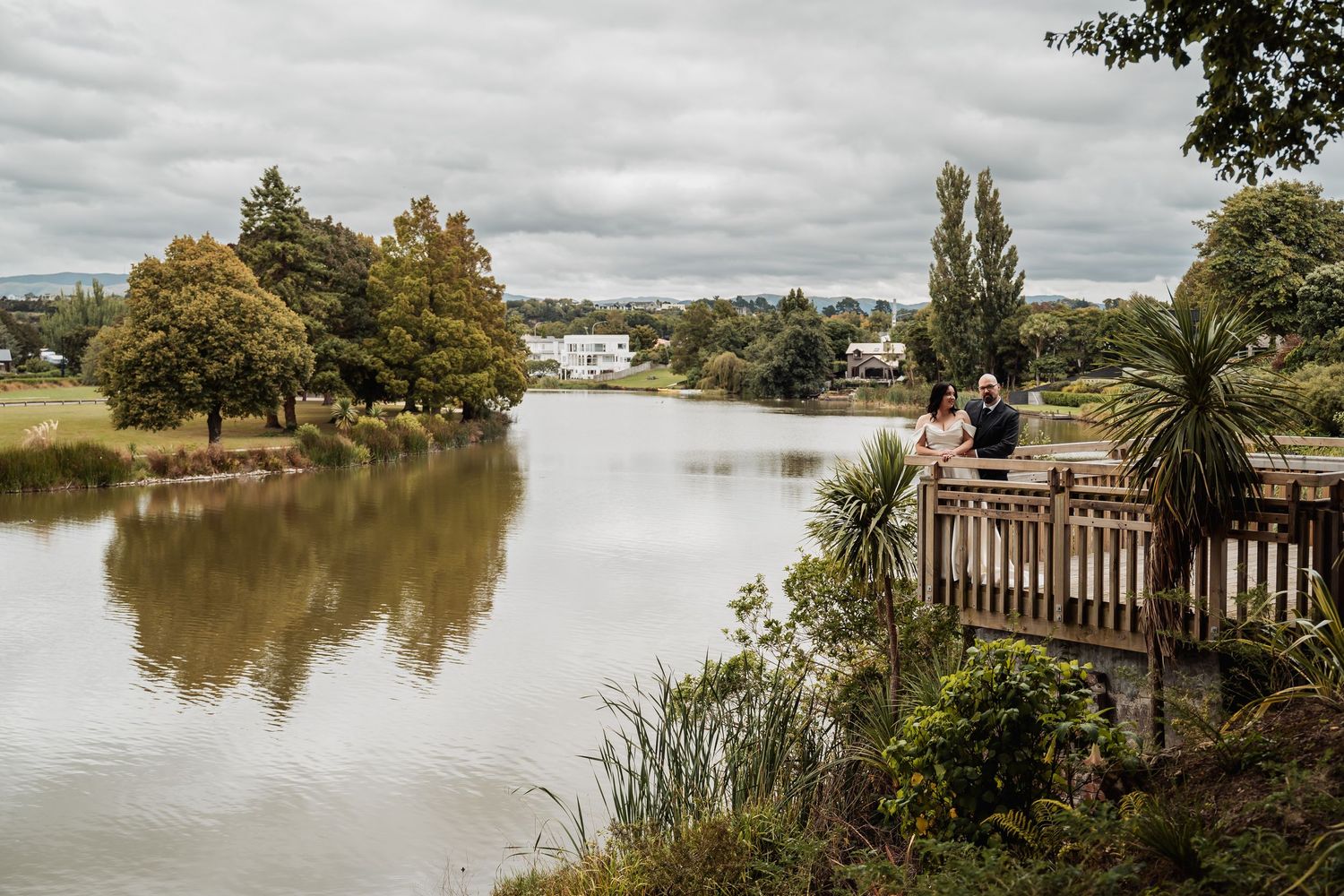 Couple standing on a wooden deck overlooking a calm river surrounded by lush trees under overcast skies.