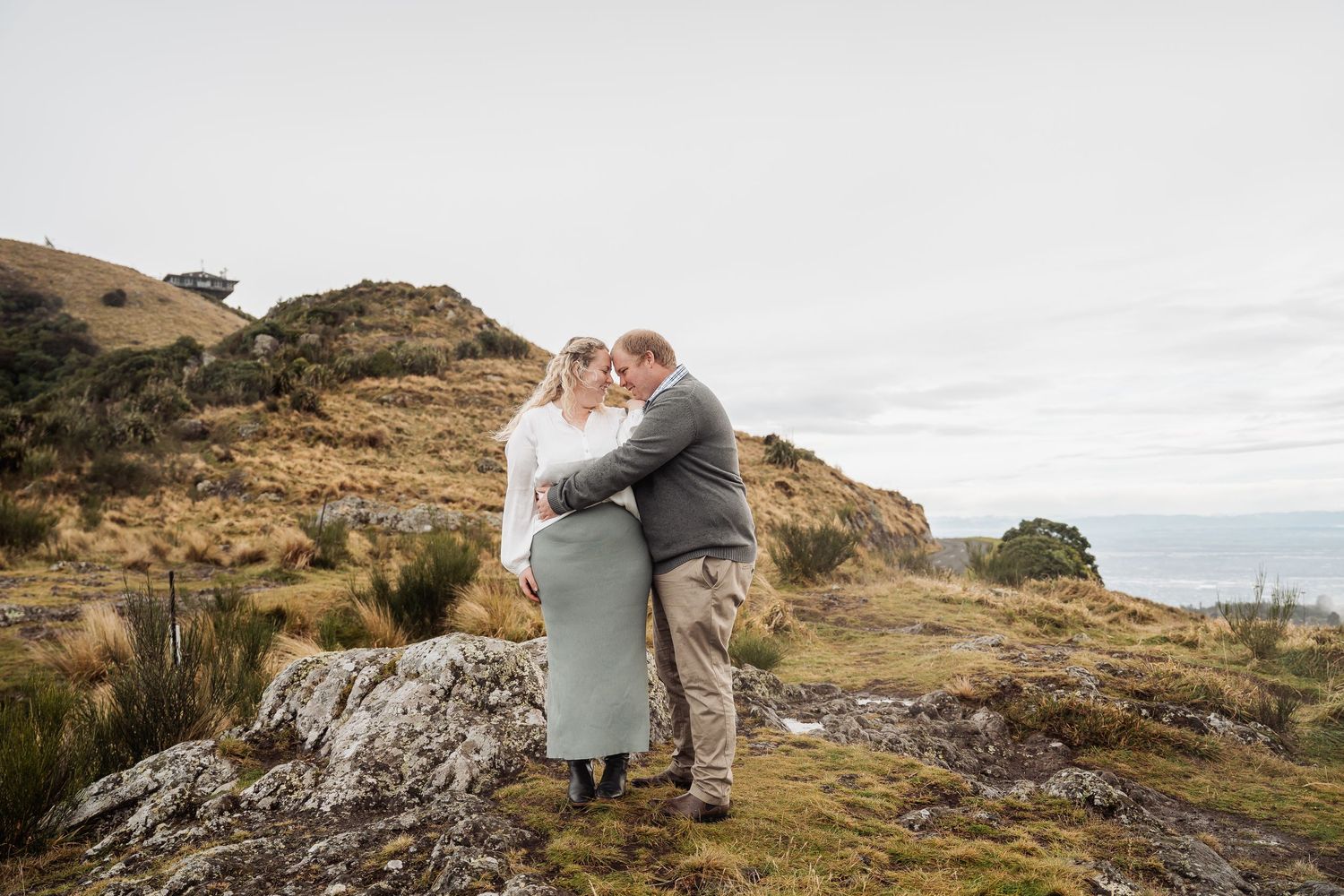 A couple embraces on a rugged coastal cliff with dramatic ocean views during sunset.