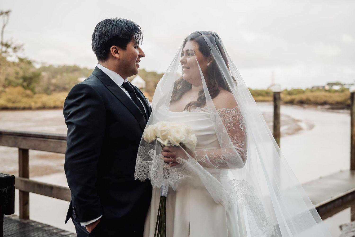 A couple in wedding attire embraces on a boardwalk with coastal scenery in the background.