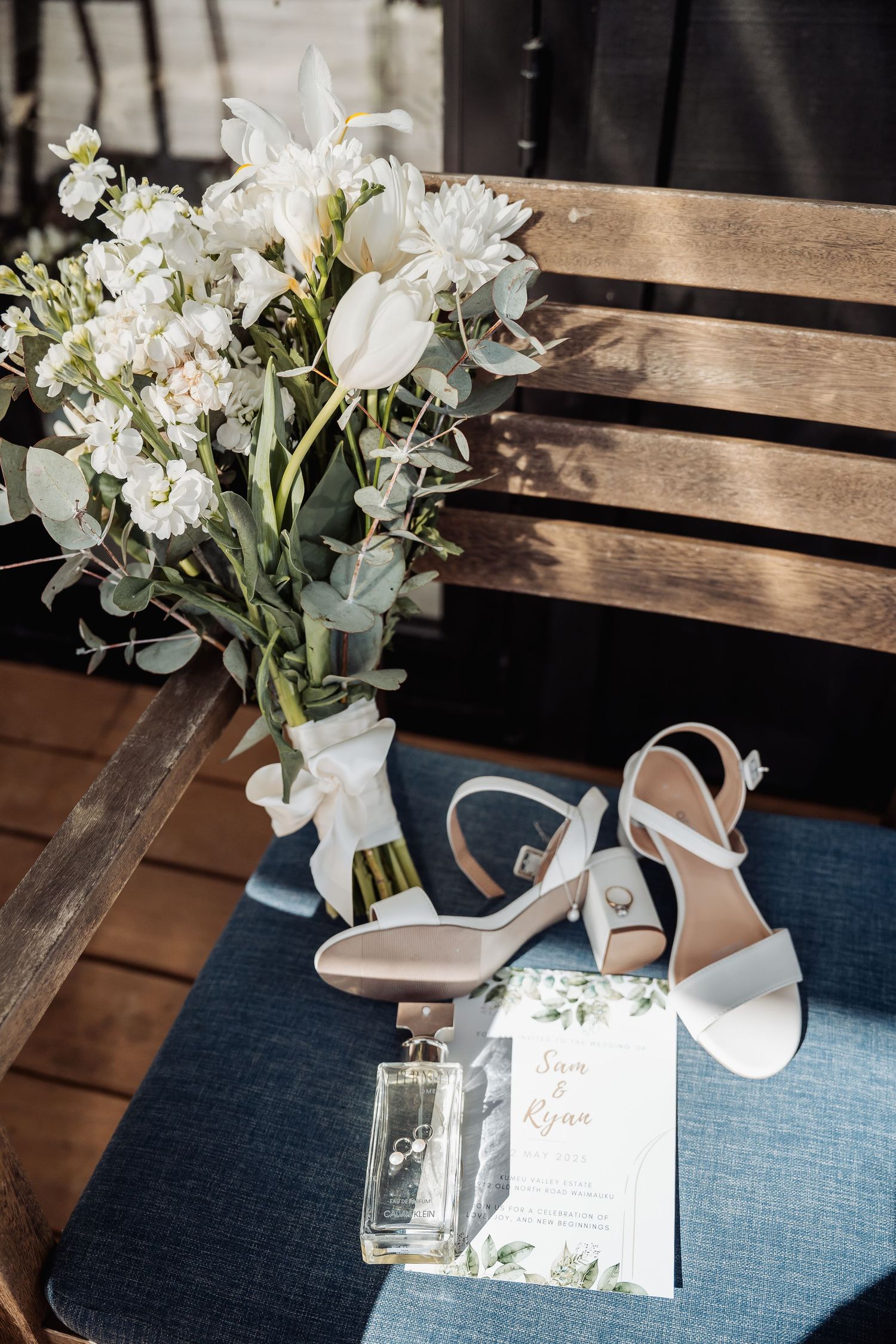 White floral bridal bouquet with elegant sandals, perfume bottle and invitation card arranged on blue cushioned bench.