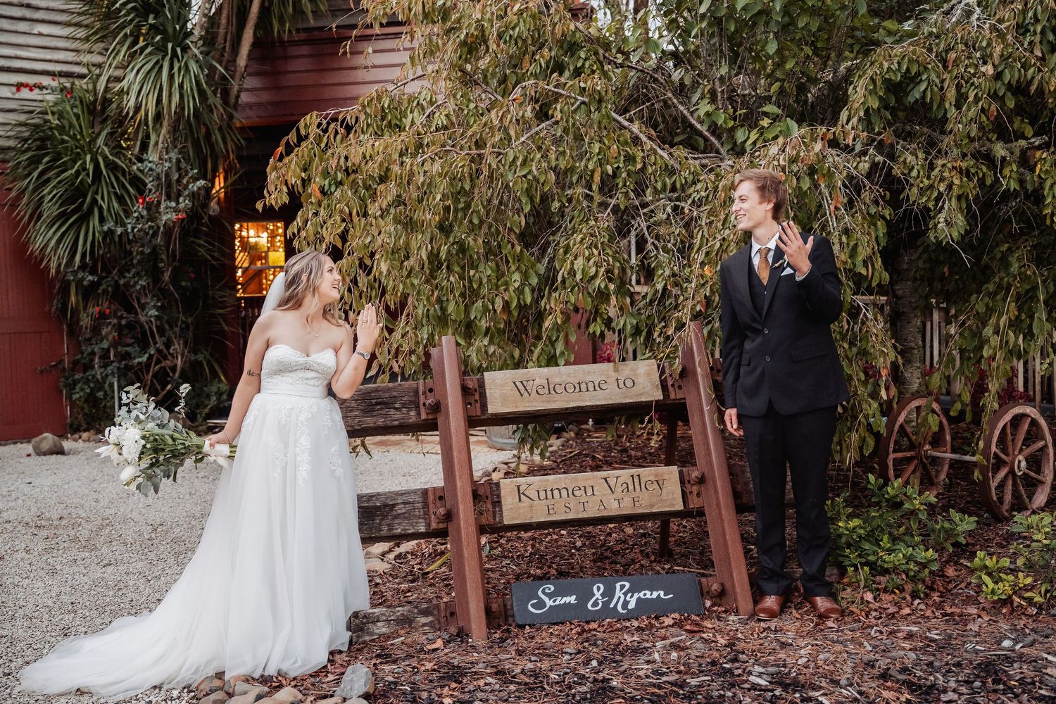 A rustic outdoor wedding scene with two people standing beside a wooden fence and palm trees at dusk.