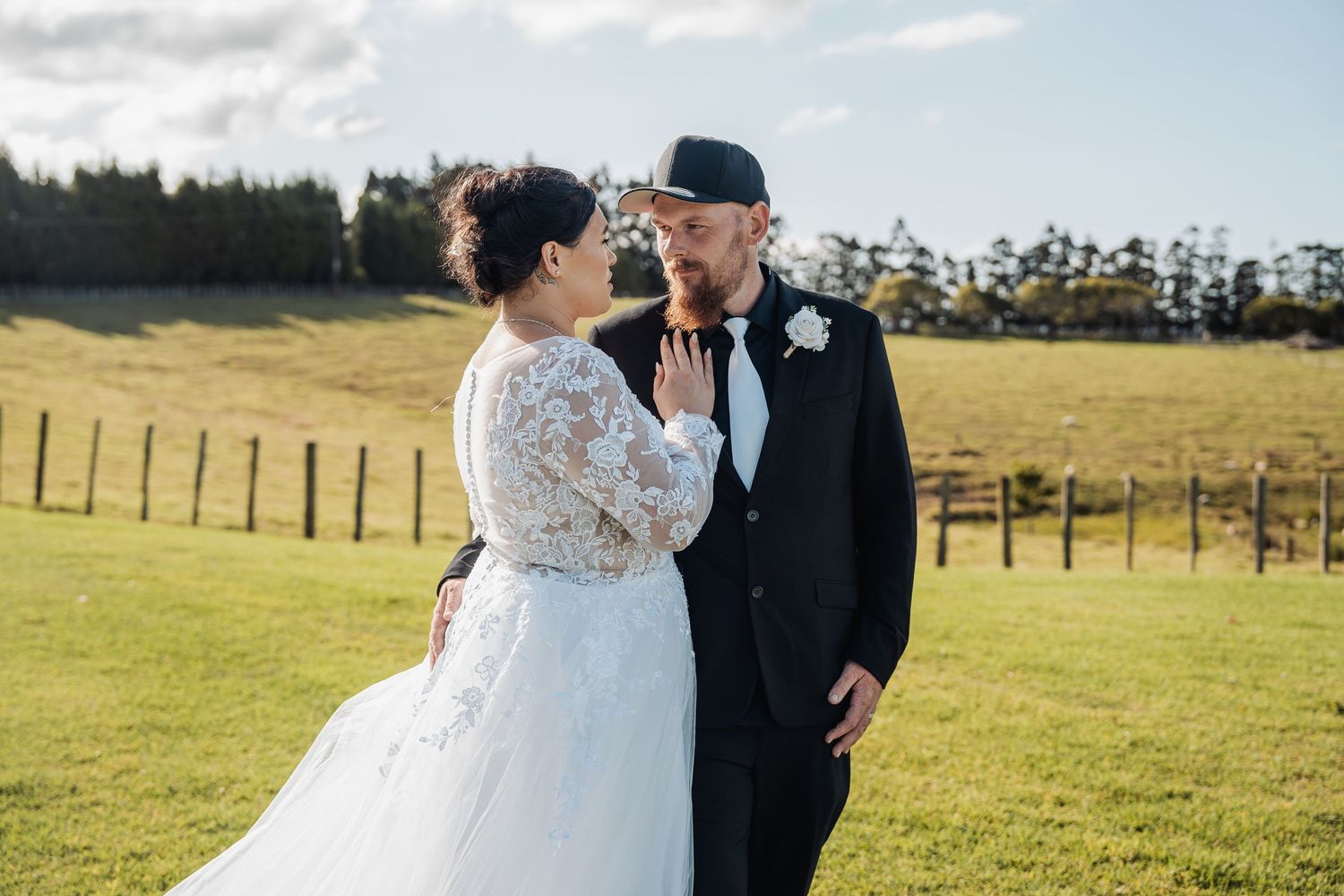 A couple shares an intimate moment on their wedding day in a sunlit green field with fencing and trees in the background.
