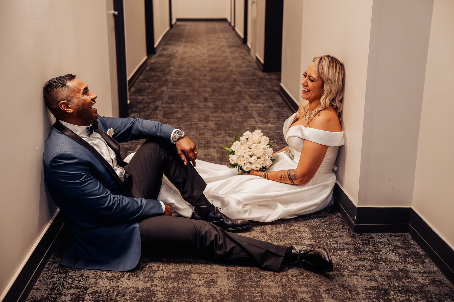 Newlyweds share a playful moment sitting on carpeted hallway floor with white bouquet.