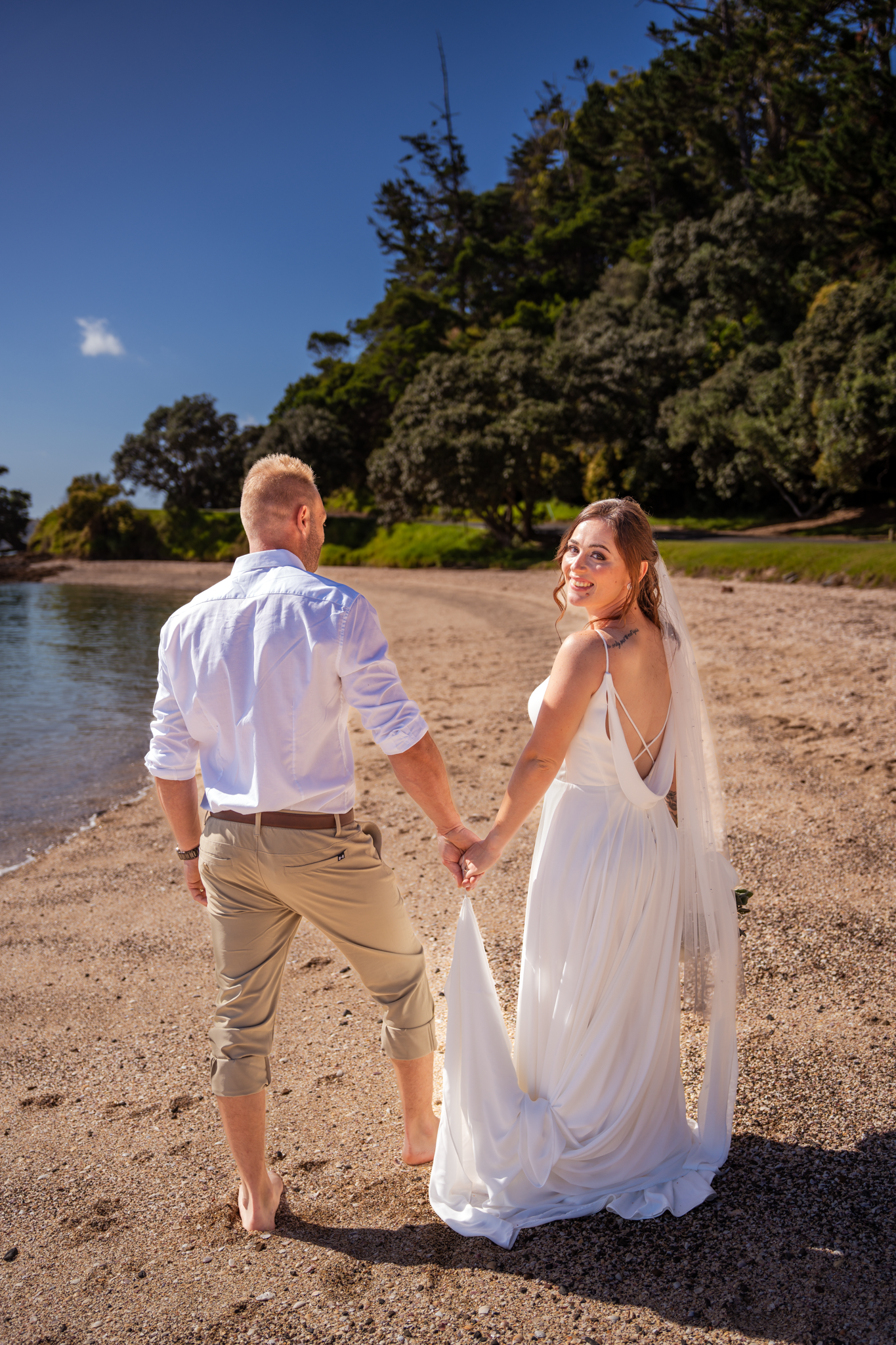 Wedding couple walks hand in hand along a scenic beach with trees in the background.