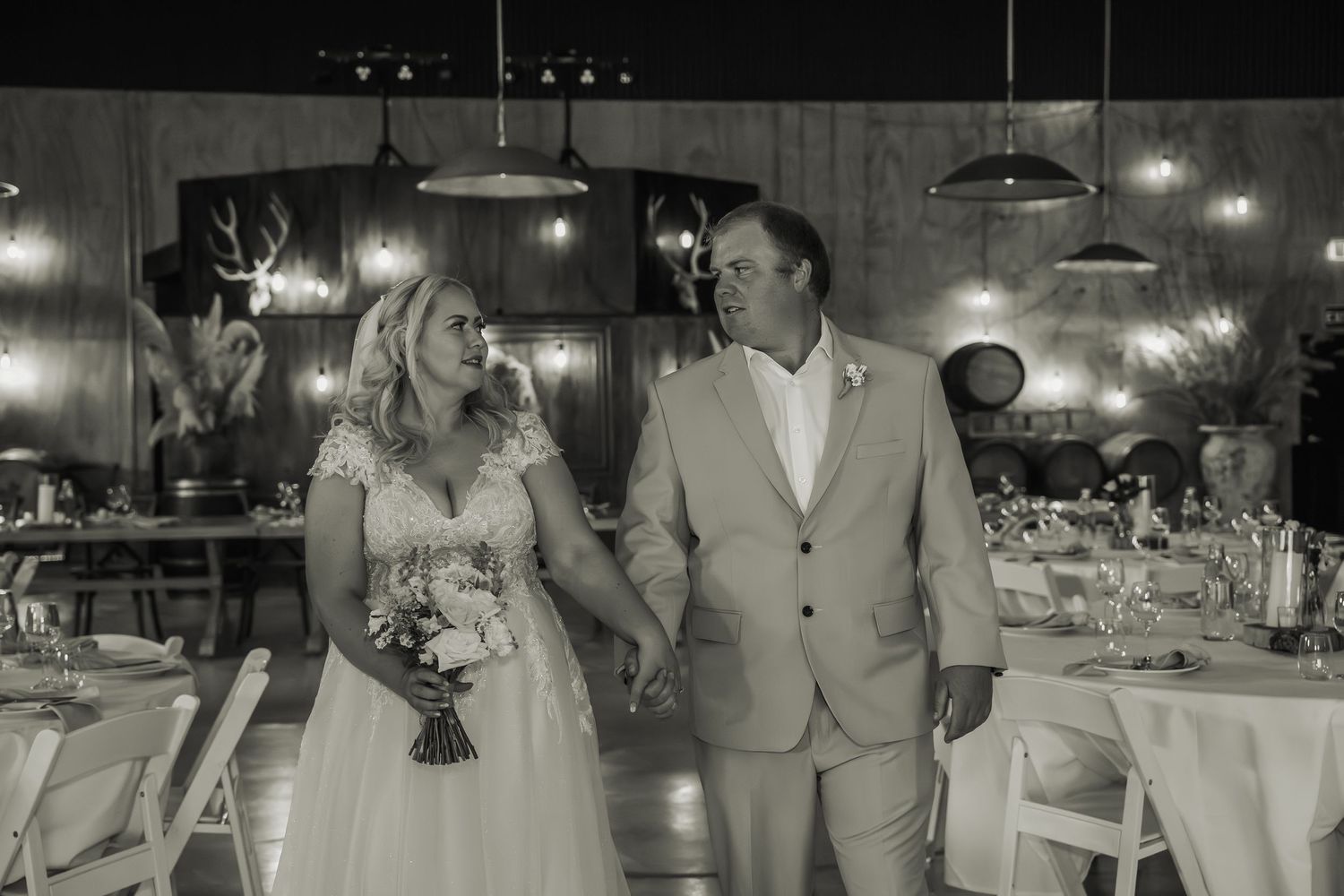 Bride and groom walk hand in hand through a warmly lit rustic reception hall in a black and white photo.