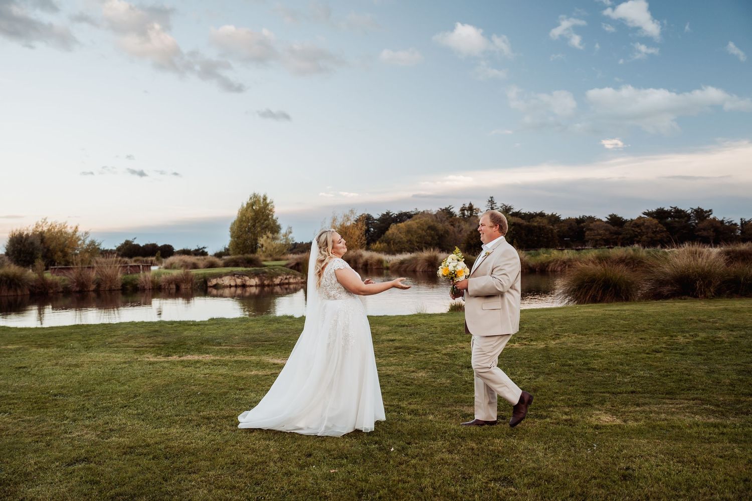 Bride and groom holding hands and dancing on green lawn beside a scenic pond.