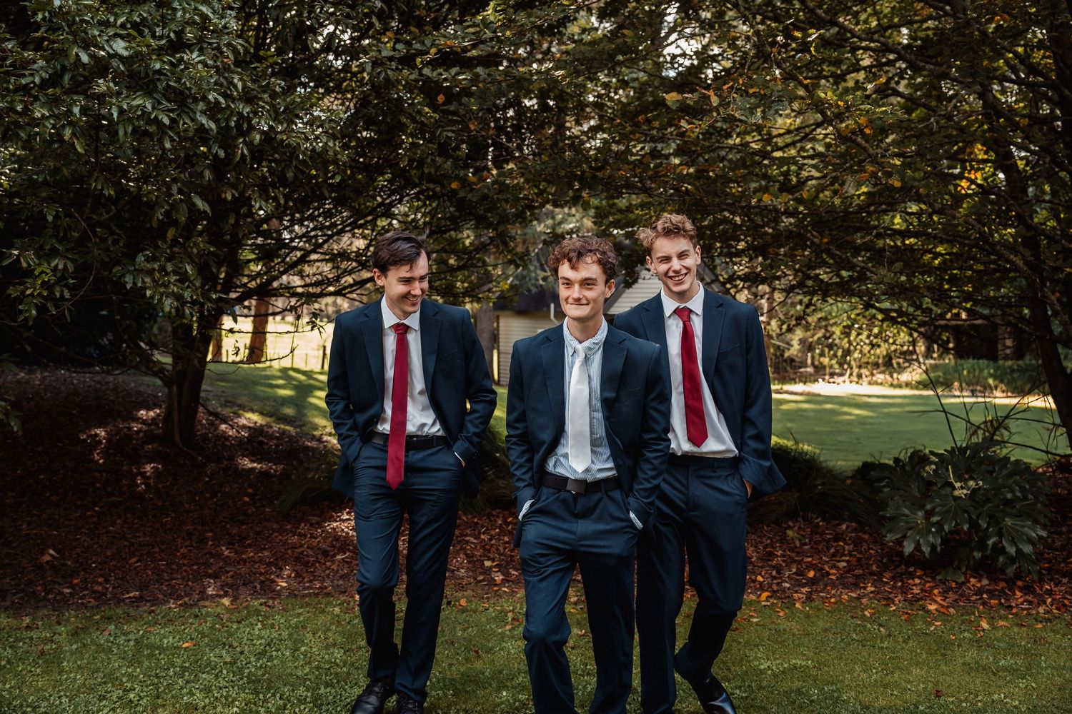 Three groomsmen in navy suits and red ties smiling outdoors among lush green trees.