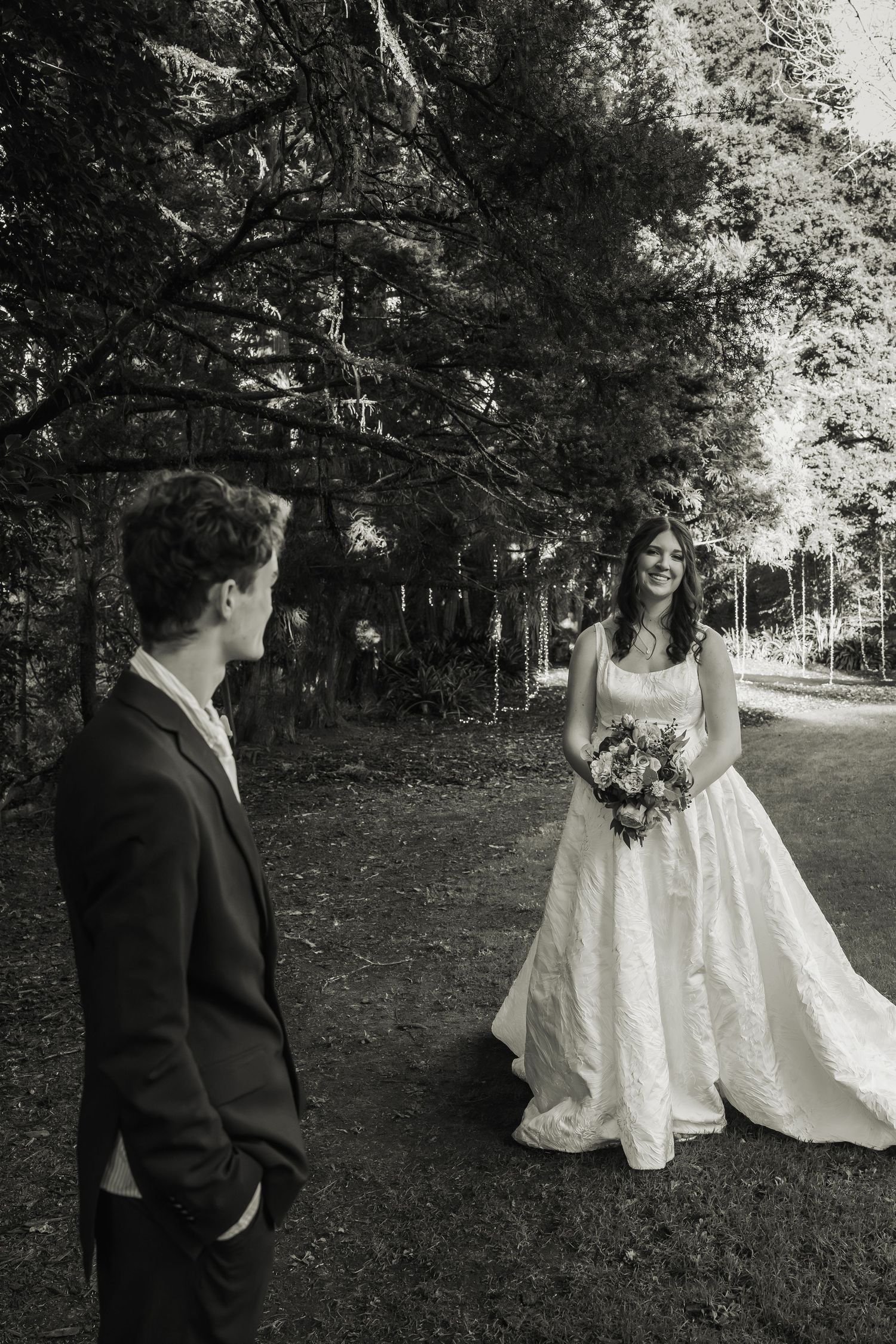 Black and white photo of groom watching bride approach in elegant wedding gown.