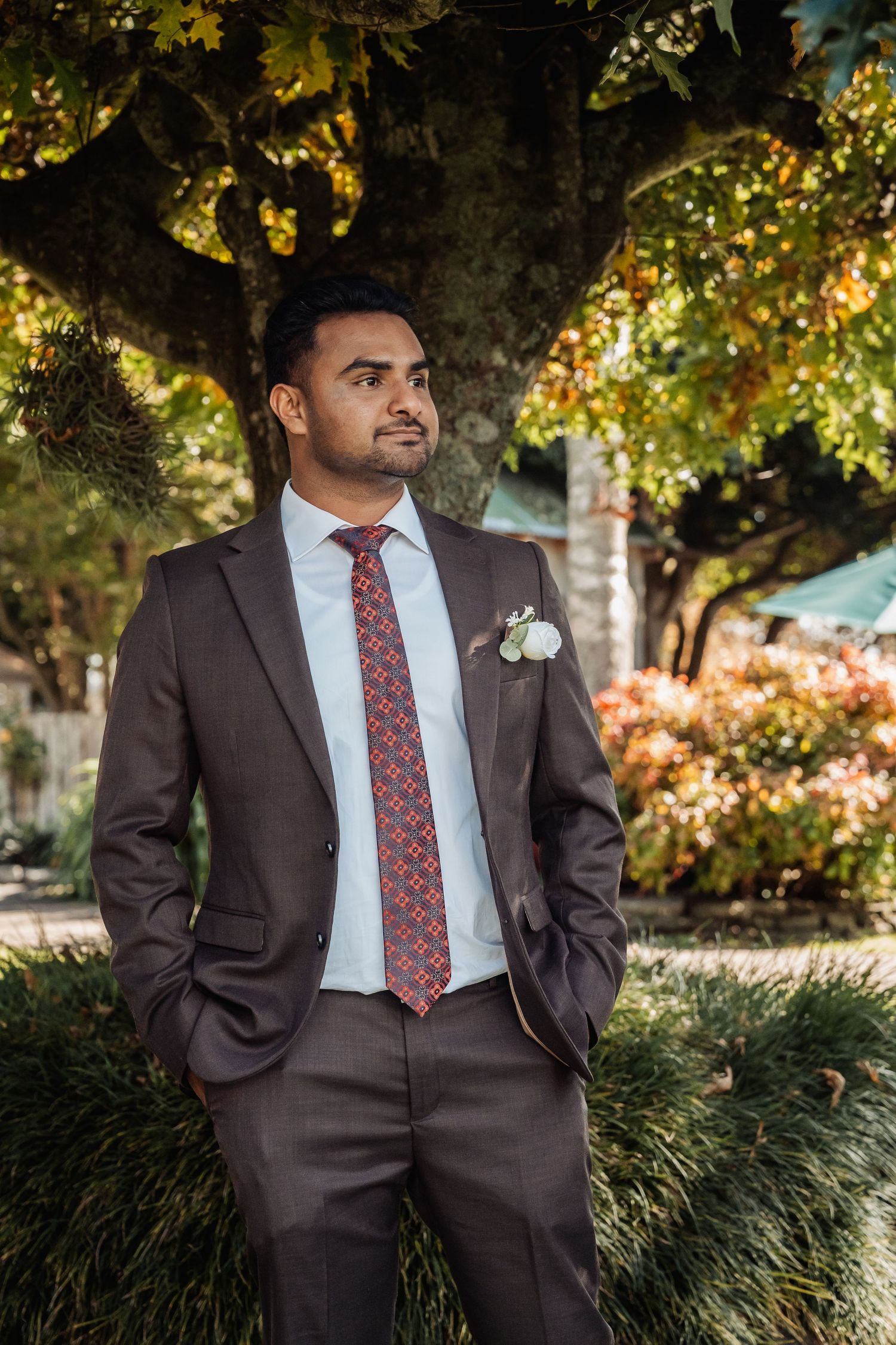 Groom in brown suit with red patterned tie and boutonniere standing outdoors among autumn foliage.