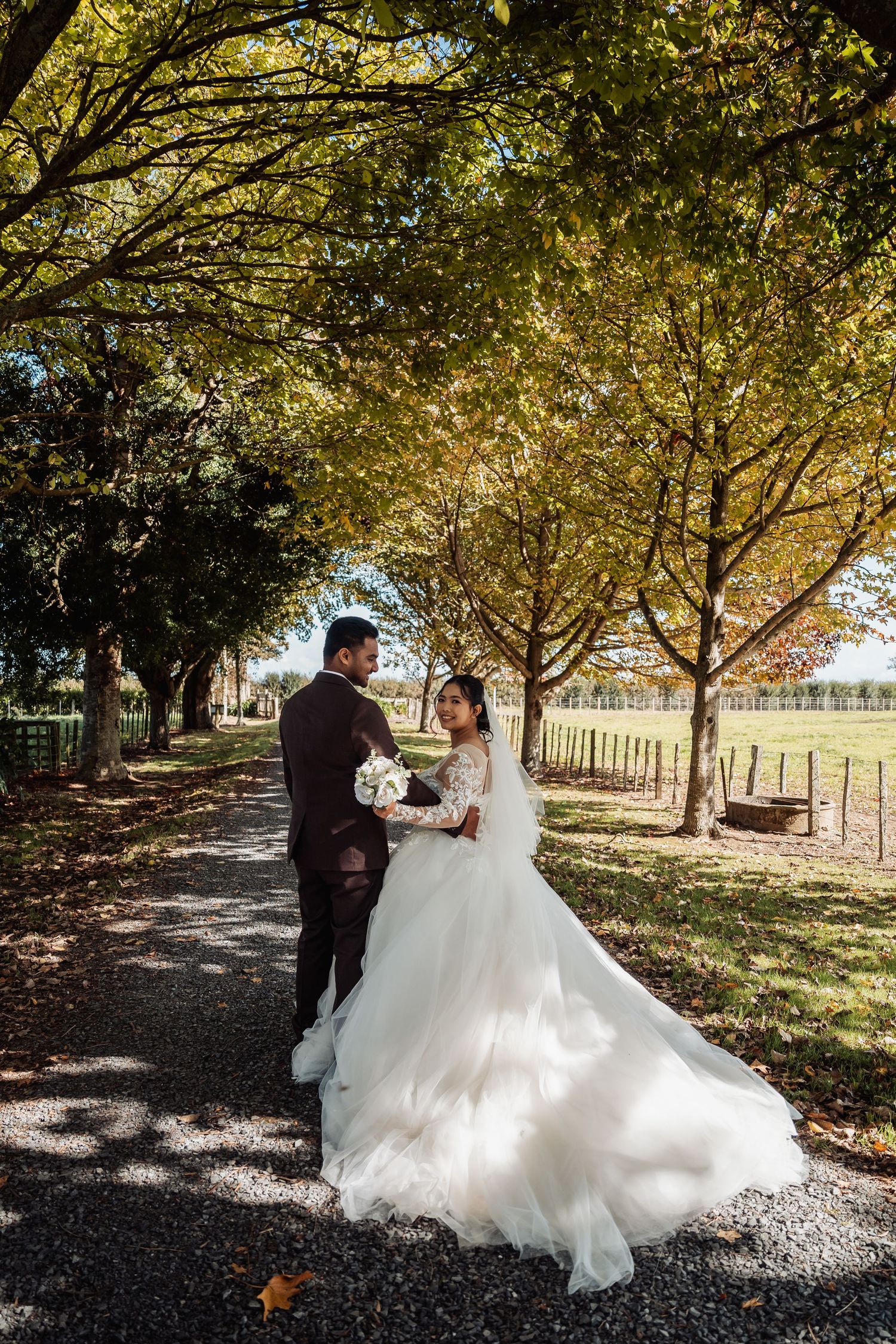 Wedding couple embracing on a sunlit tree-lined path, bride in flowing white gown holding a white floral bouquet.