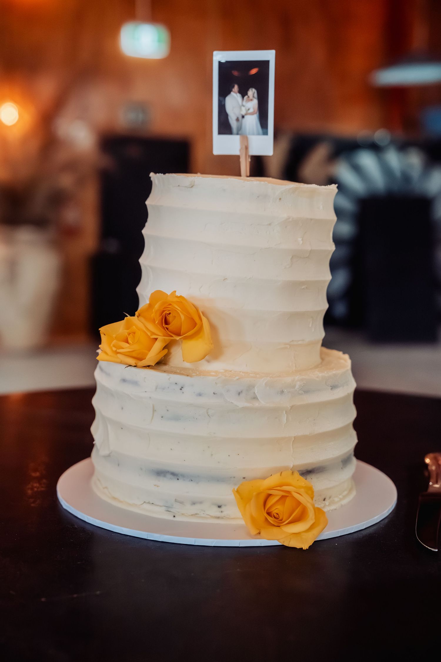 Two-tier white wedding cake with yellow roses and Polaroid photo topper on dark table.