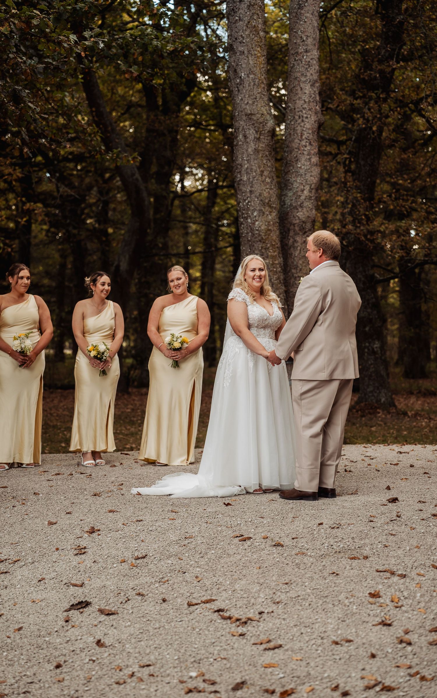 A bride in a white gown exchanges vows with her groom as bridesmaids in yellow dresses stand beside her outdoors.