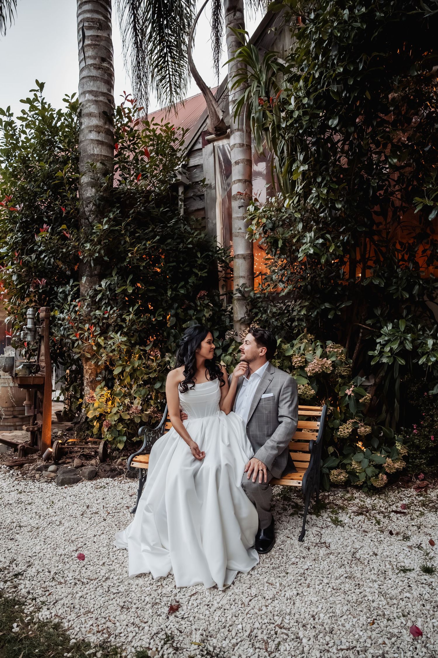 Bride and groom sit together on a bench surrounded by lush tropical greenery during outdoor wedding portrait session.