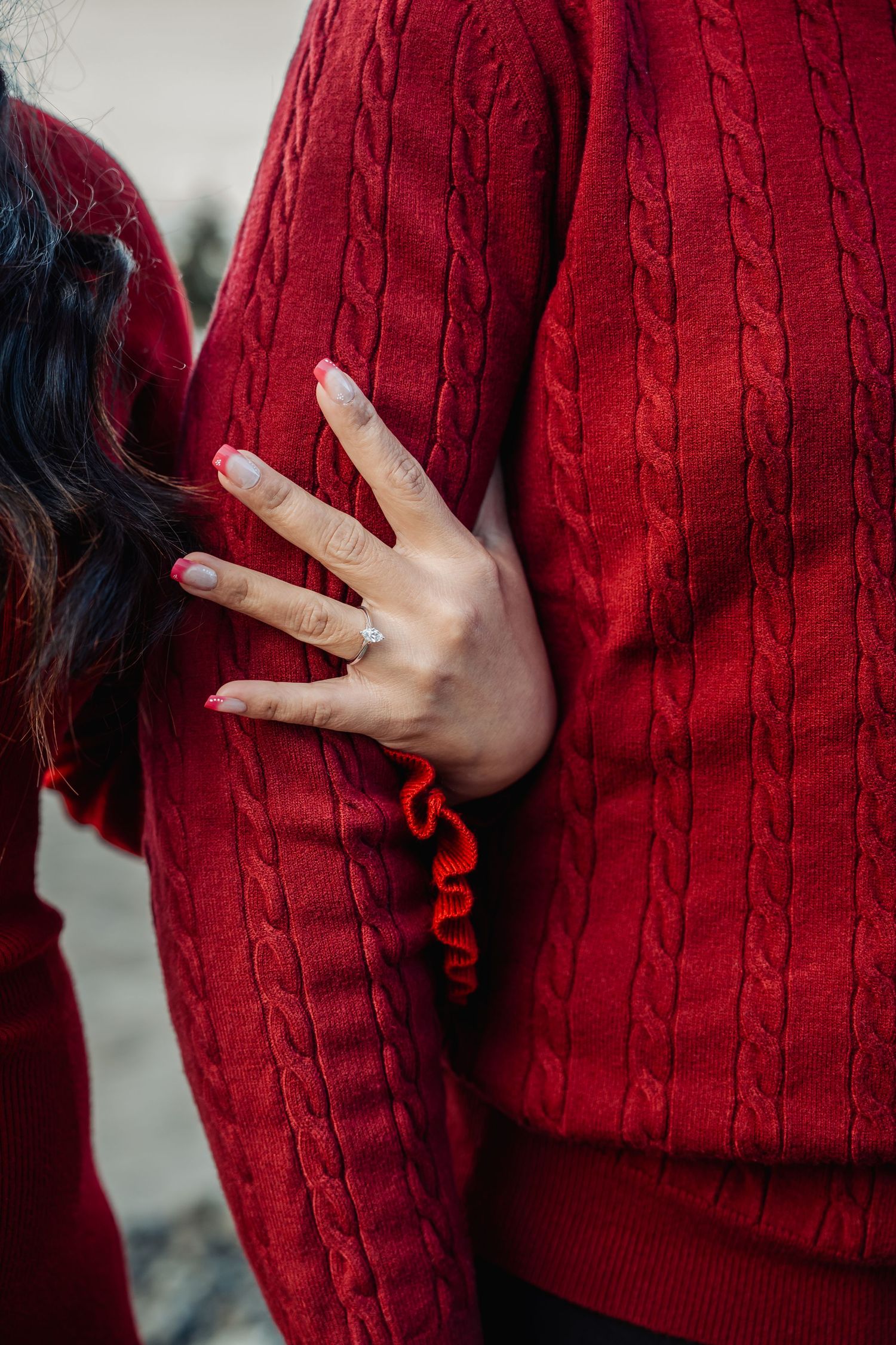 A woman displays her engagement ring while embracing a man, both wearing matching red cable-knit sweaters.