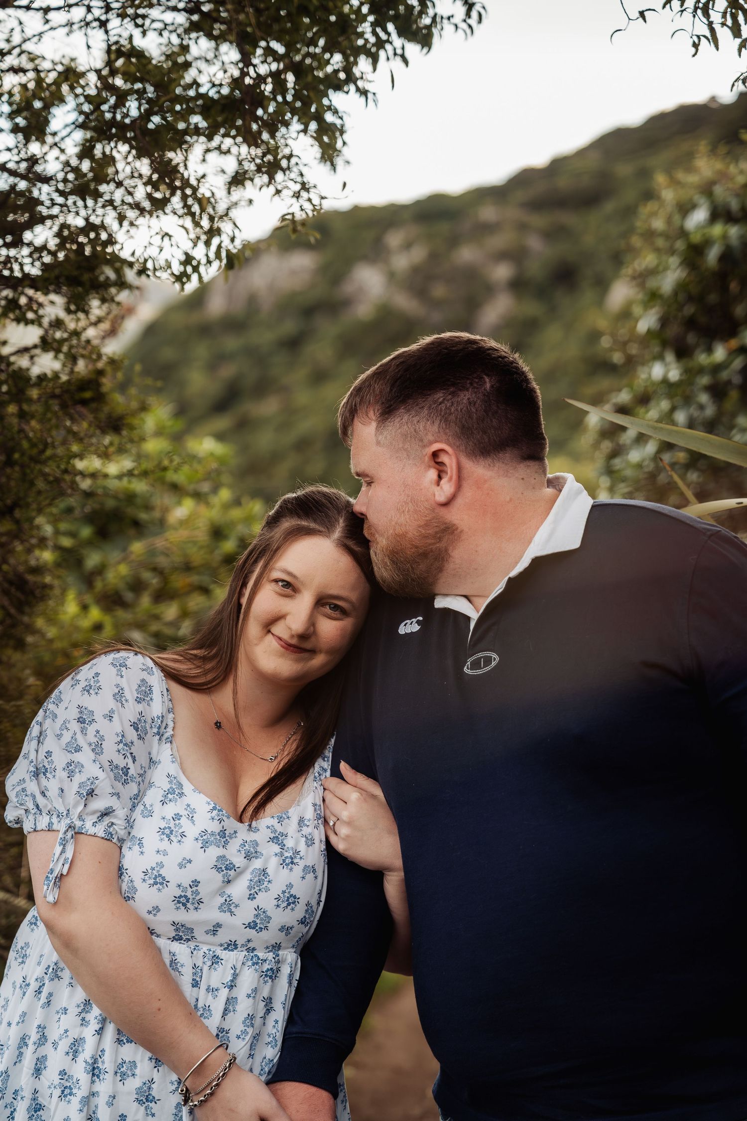 Man kissing woman on cheek as she smiles softly, standing together in a lush green nature setting.