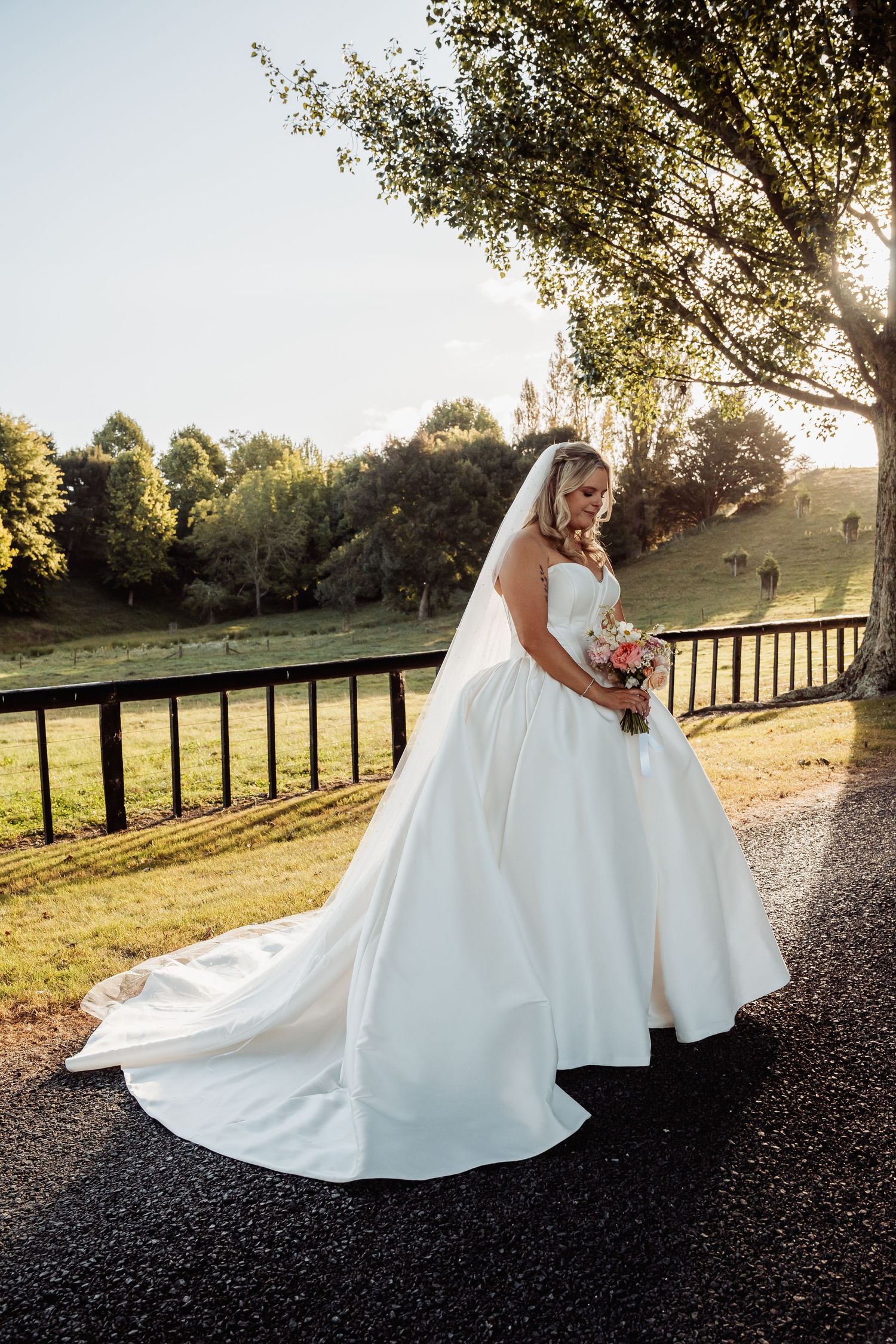 Elegant bride in strapless ball gown with long train stands on sunlit path holding pink bouquet.