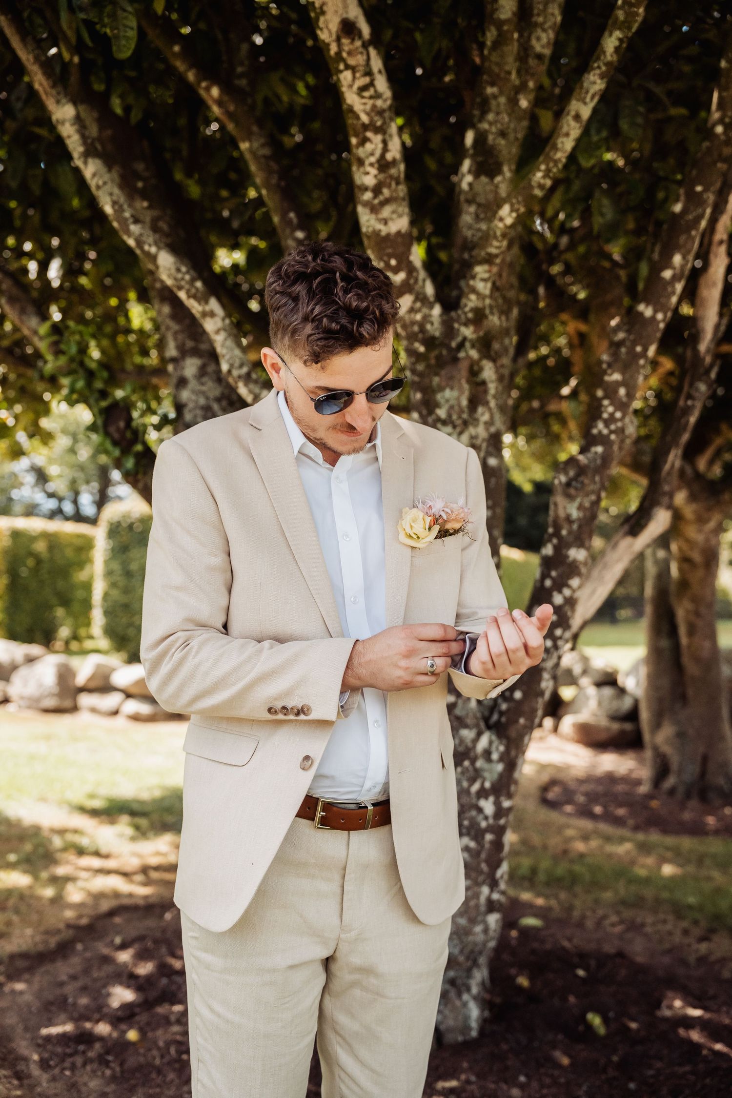 Groom in beige suit adjusting cufflinks outdoors near trees.
