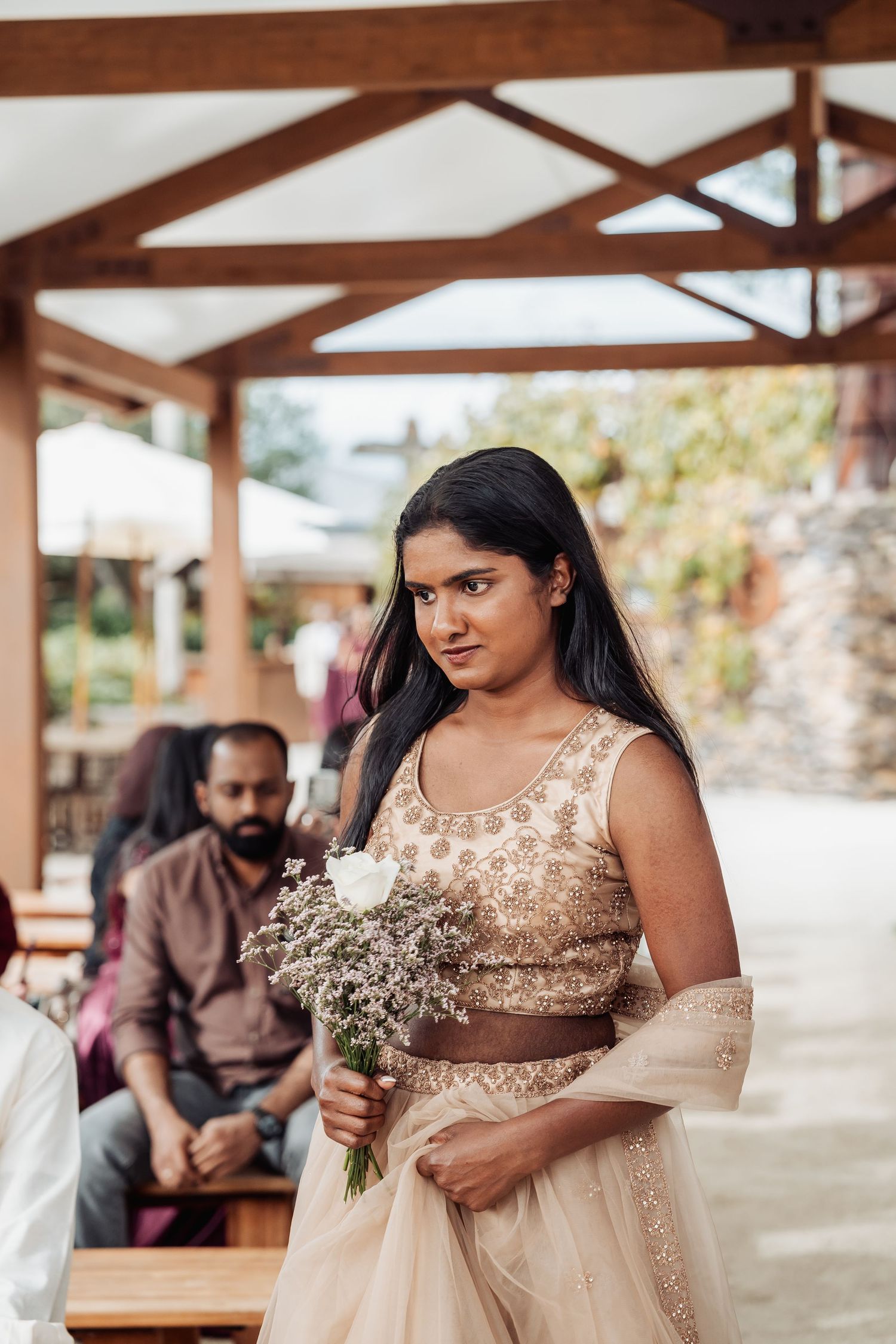 Bridesmaid in gold lehenga holding white rose bouquet during outdoor wedding ceremony.