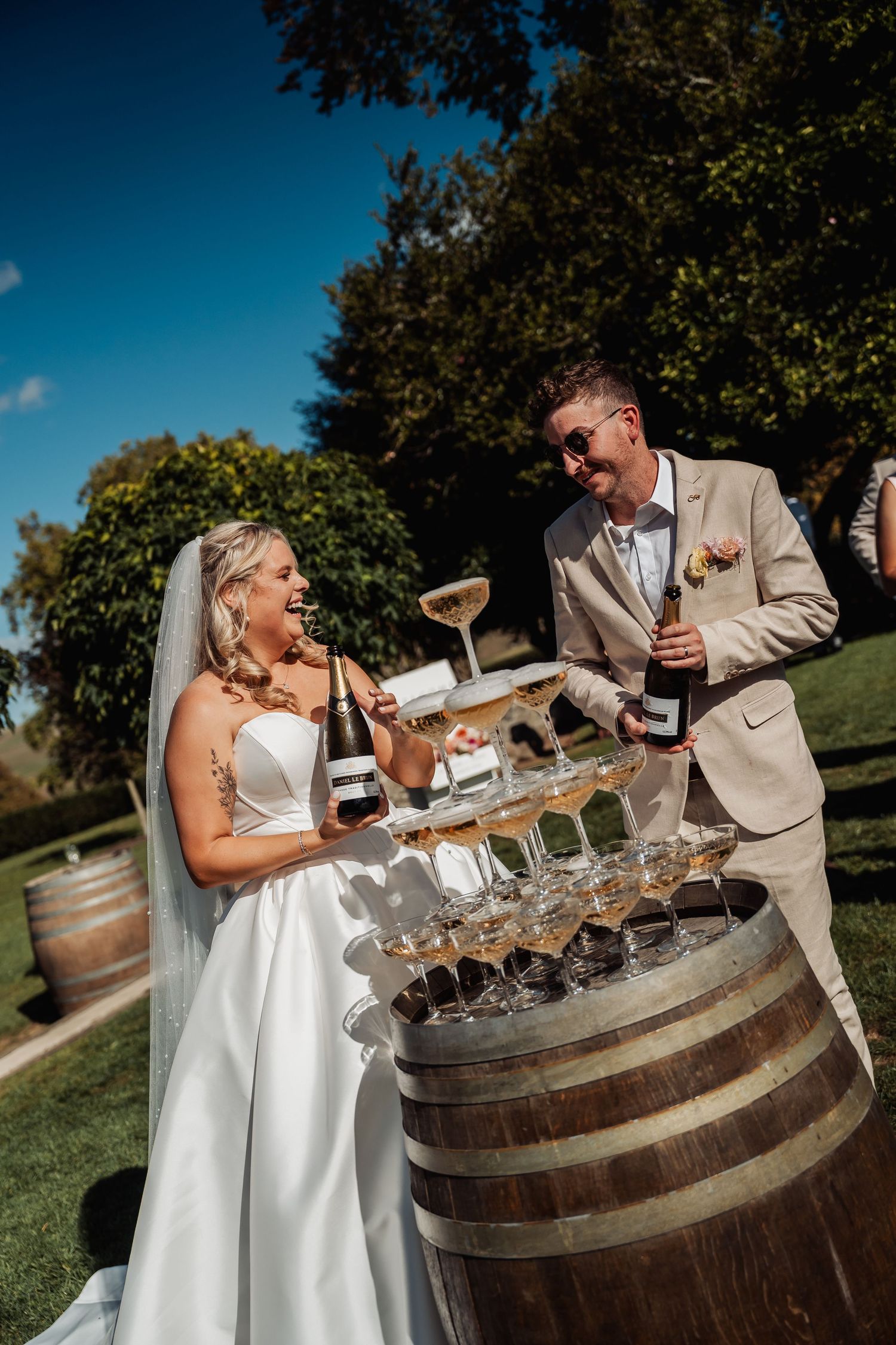 Joyful newlyweds pouring champagne tower on a wooden barrel at their sunny outdoor wedding.