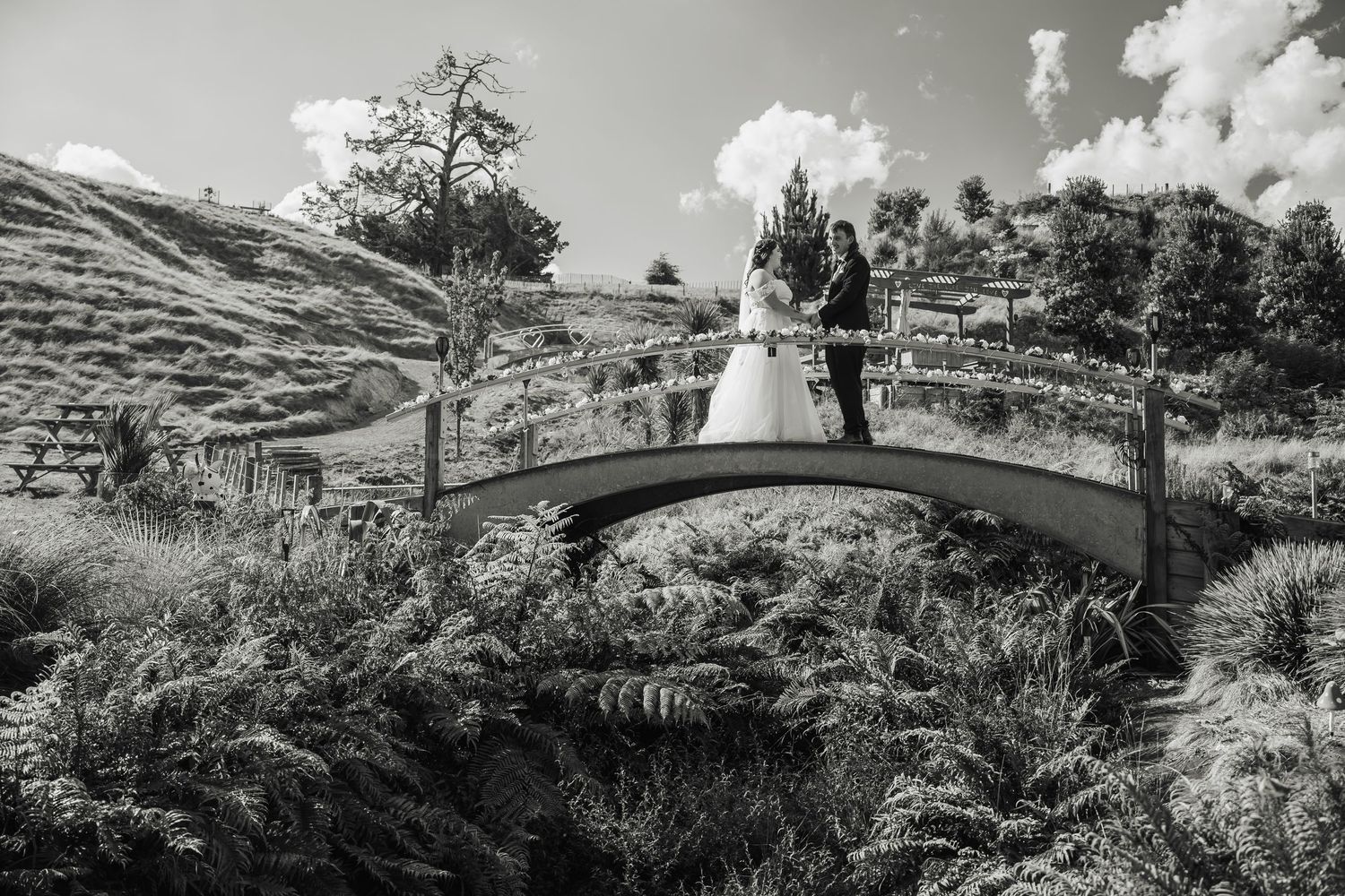 Bride and groom standing on a charming garden bridge surrounded by lush greenery in black and white.