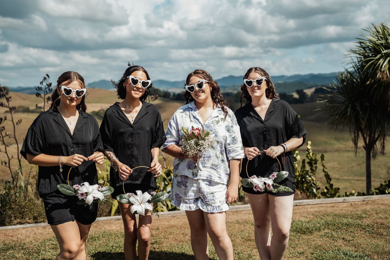 Four bridesmaids in black outfits and heart sunglasses walking outdoors holding floral arrangements.