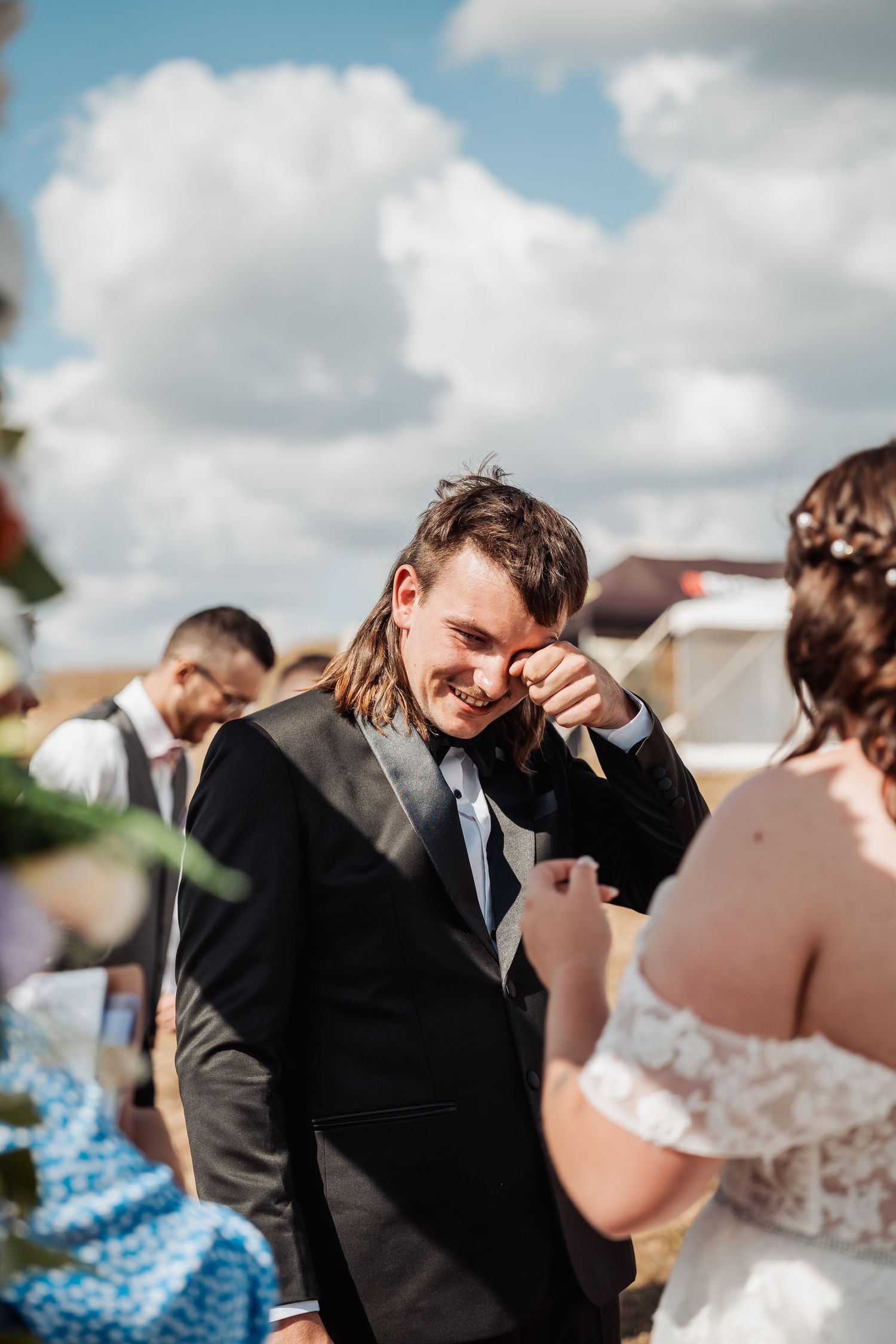 Emotional groom in black suit wipes tear while smiling at bride during outdoor wedding ceremony.