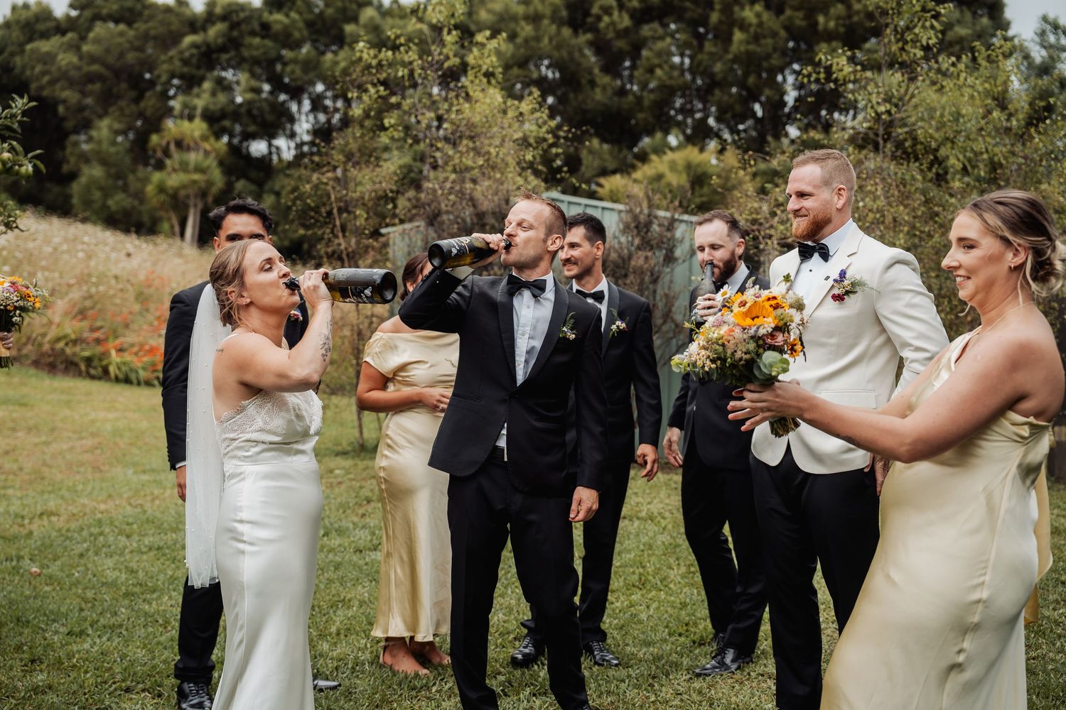 Wedding party celebrating outdoors with bride and groom drinking from bottles with bridesmaids and groomsmen.