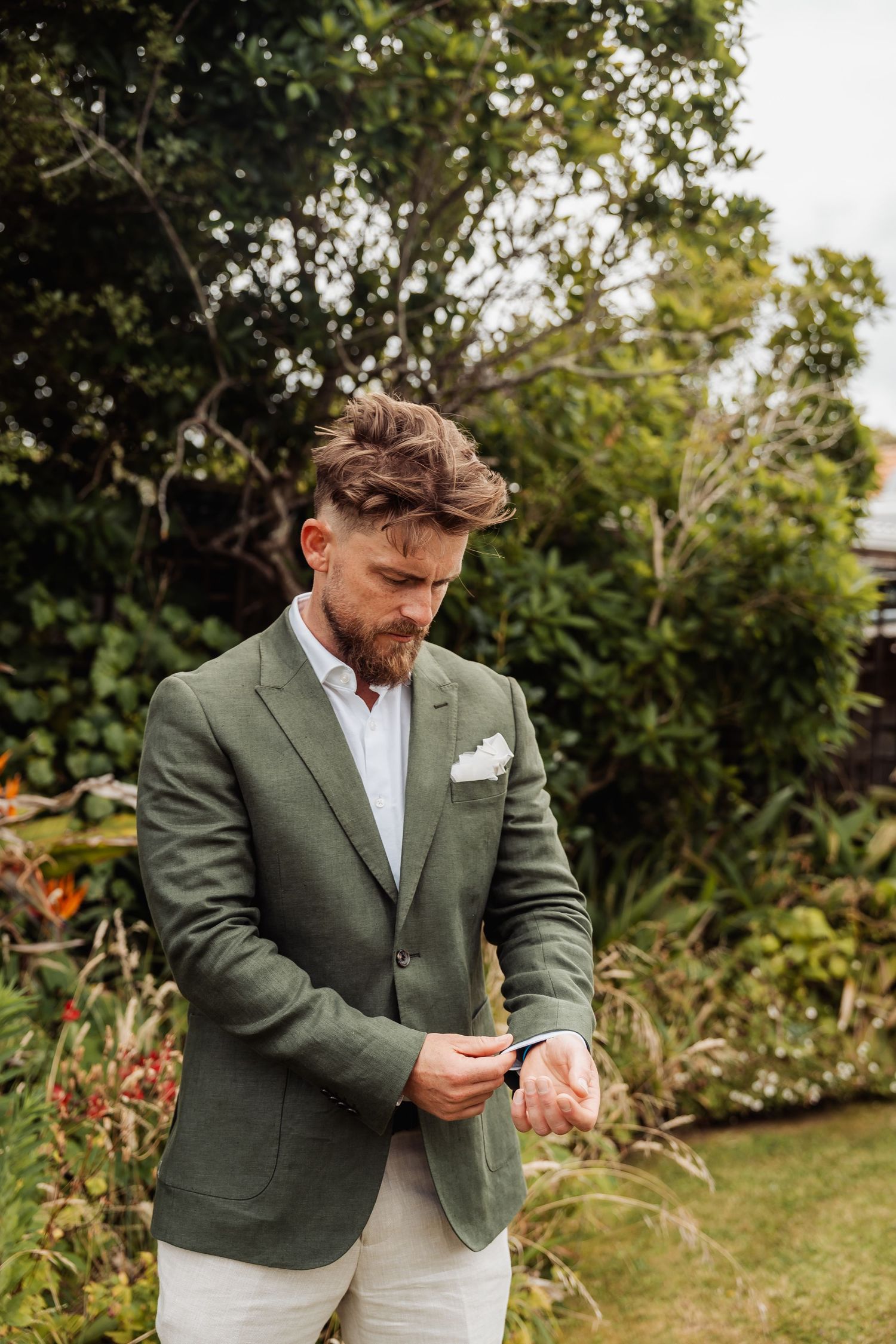 Stylish groom in green blazer and white pocket square adjusting his cufflinks in a garden.