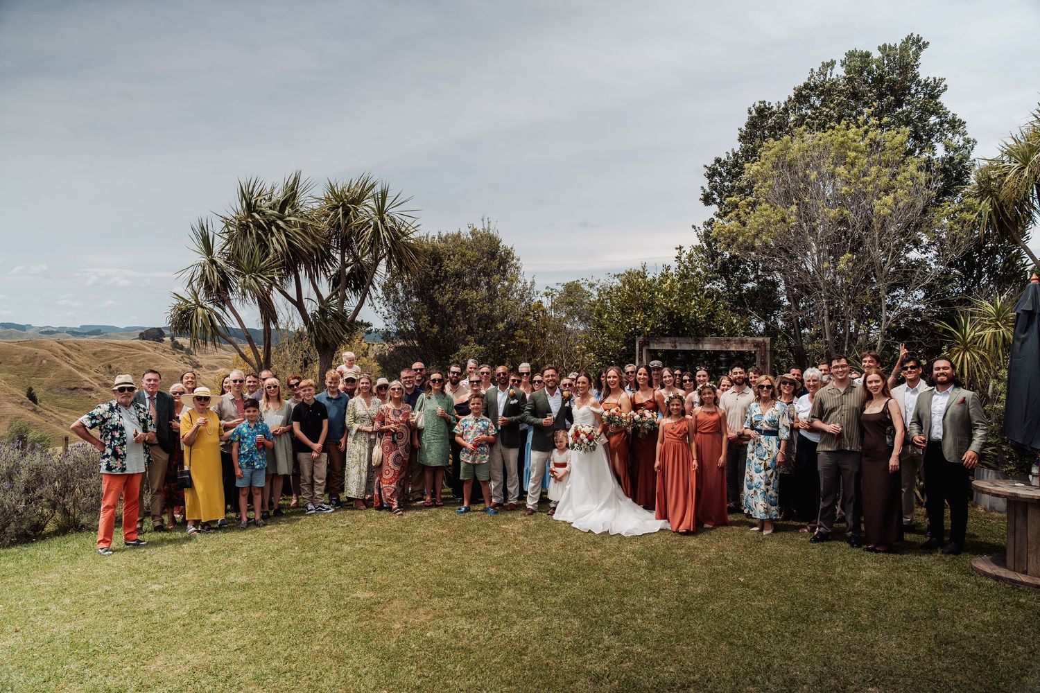 Large wedding group photo with bride and groom surrounded by guests on a sunny outdoor lawn.