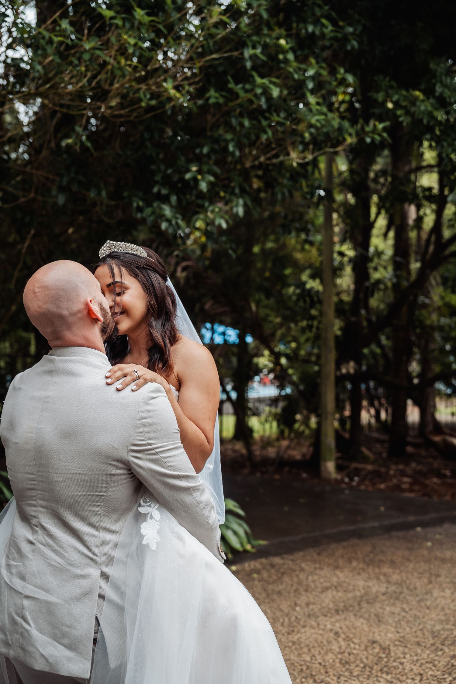 A bride and groom share a romantic almost-kiss outdoors, surrounded by lush green trees on their wedding day.